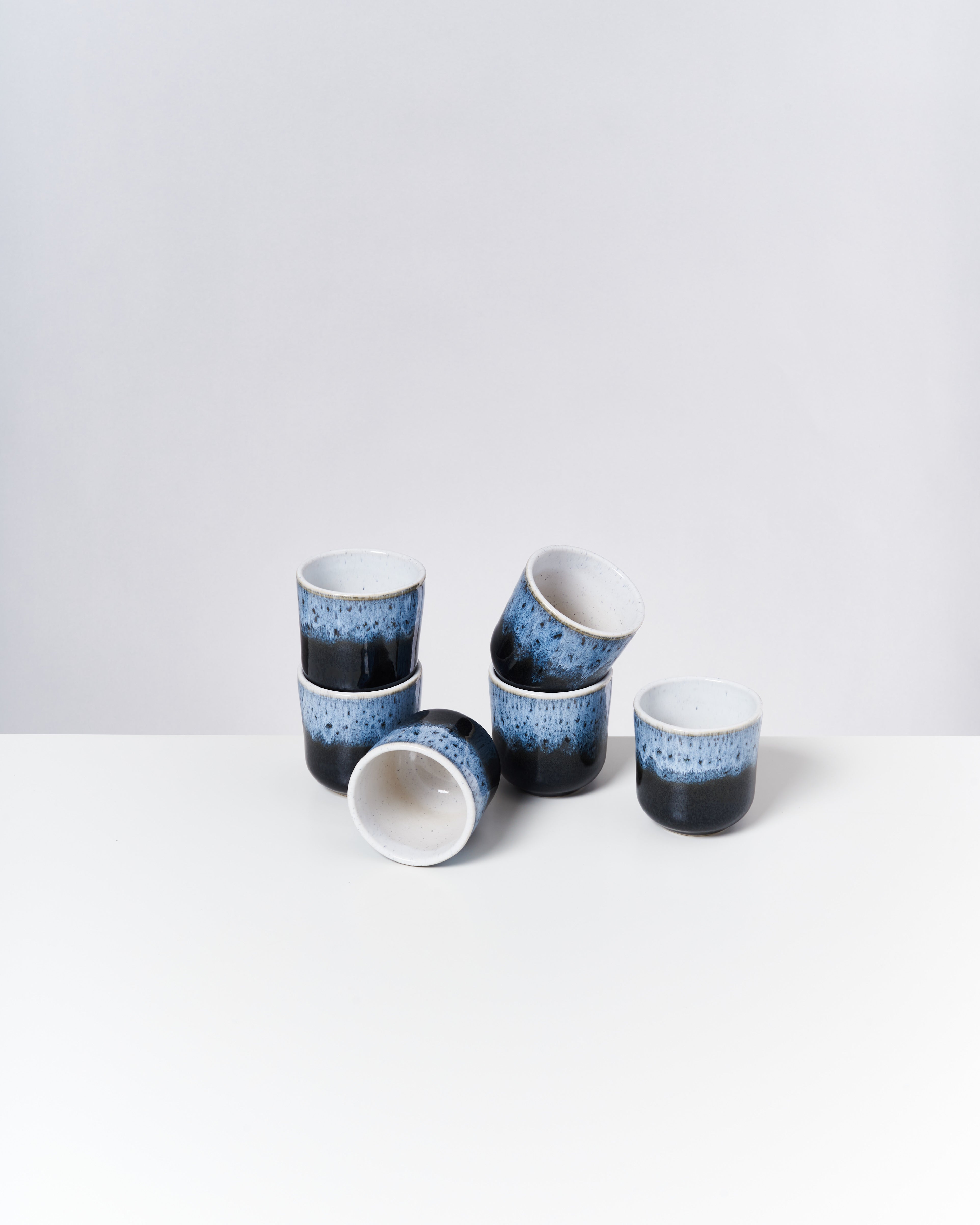 Sechs blaue Keramikbecher mit schwarzem Rand, unregelmäßig gestapelt, auf einer weißen Oberfläche vor weißem Hintergrund.