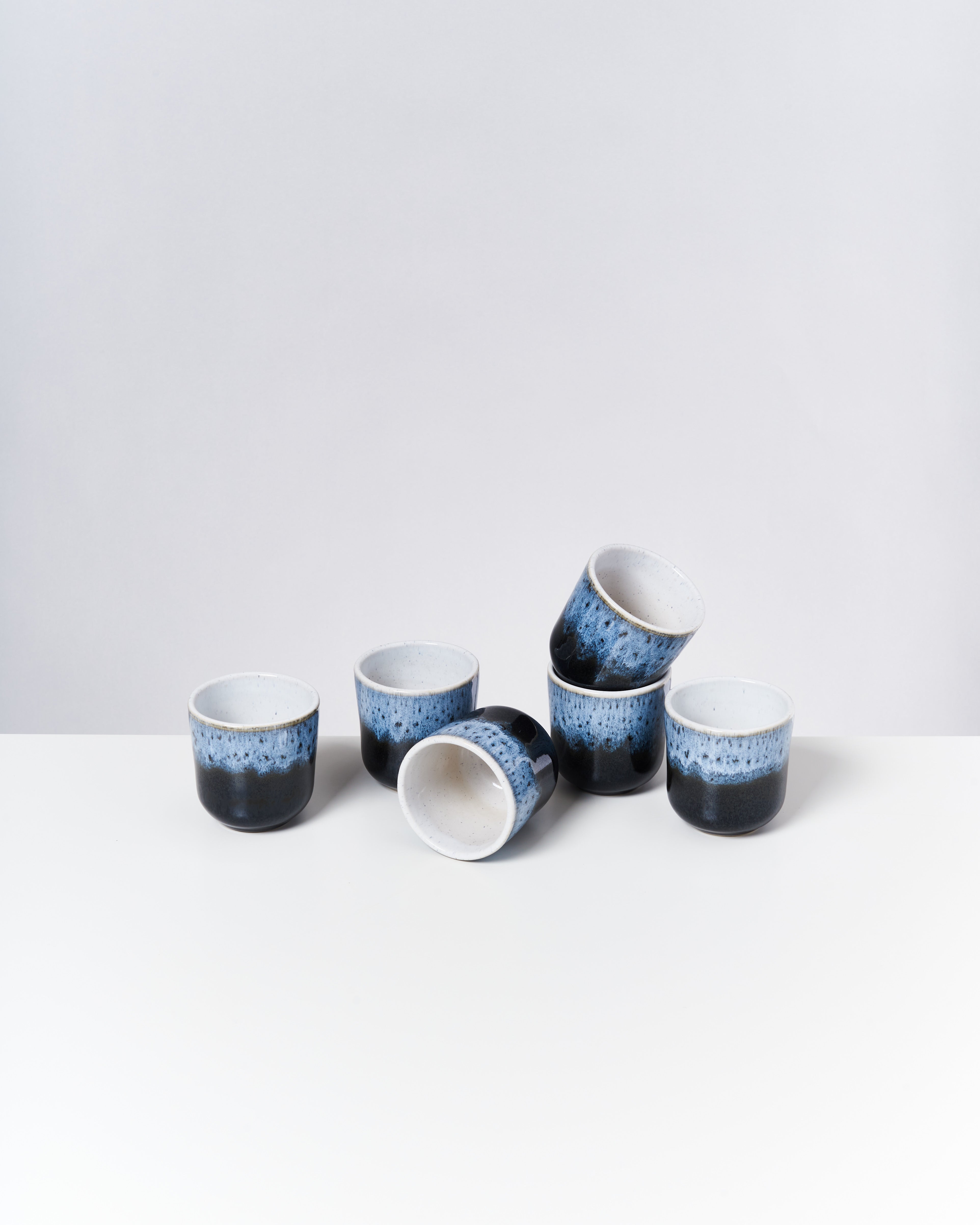 Sechs blaue Keramikbecher mit weißem Inneren stehen und liegen auf einer glatten, weißen Oberfläche.
