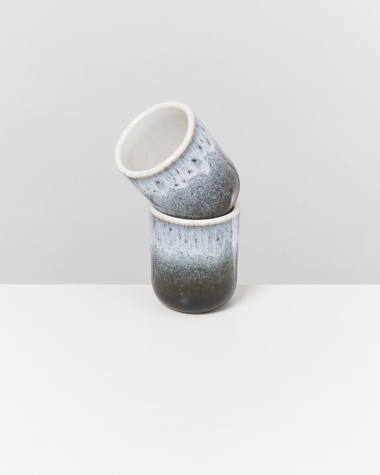 Zwei gestapelte Keramiktassen mit blauer und schwarzer Glasur auf einer weißen Oberfläche vor grauem Hintergrund.