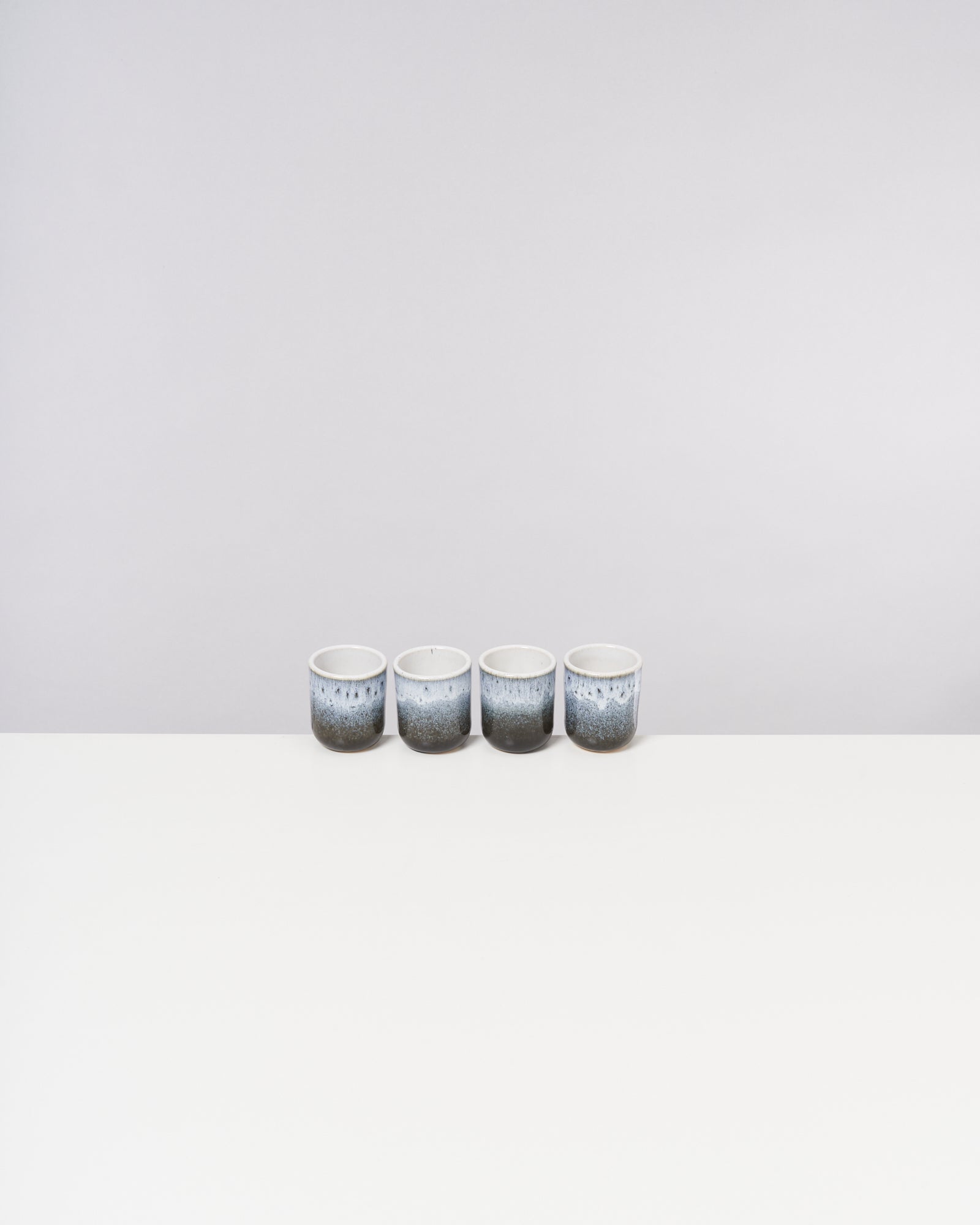 Vier Keramikbecher mit blauem Muster stehen in einer Reihe auf einem weißen Tisch vor neutralem Hintergrund.