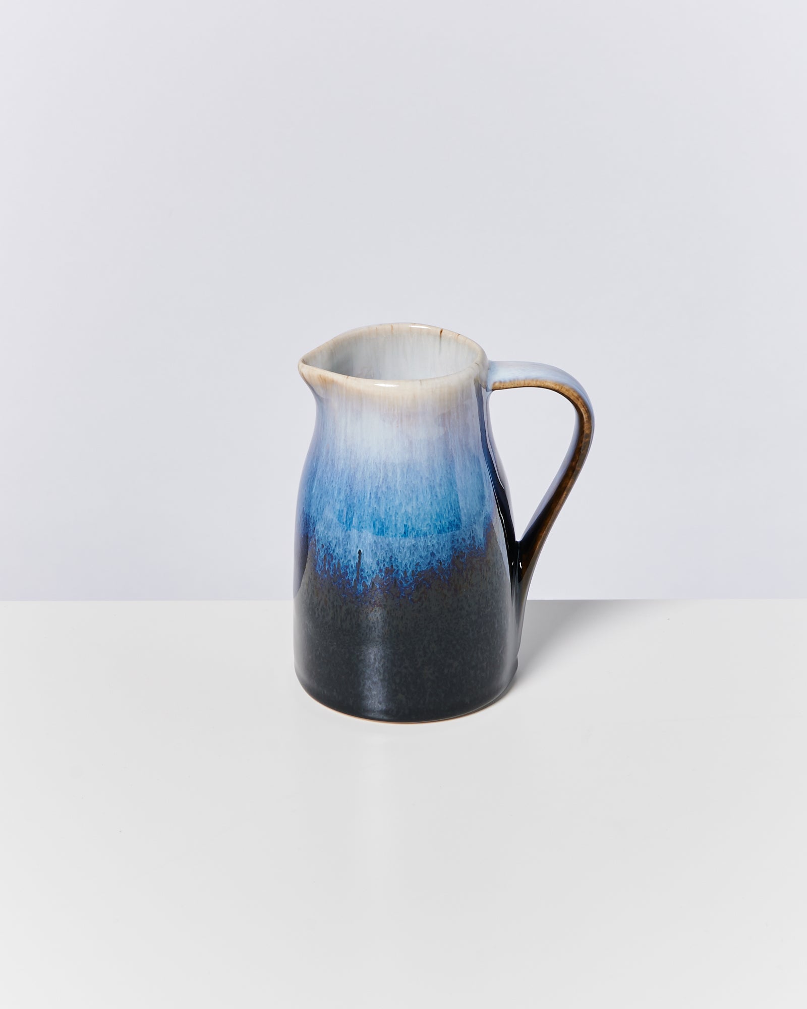 Keramikkrug mit blau-schwarzem Farbverlauf steht auf weißem Tisch vor grauem Hintergrund.