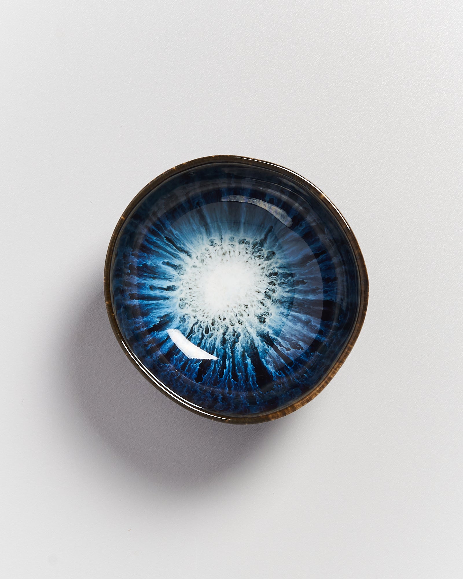 Keramikschale mit blau-weißem Muster, das von der Mitte ausstrahlt und einem Auge ähnelt, auf hellem Hintergrund.