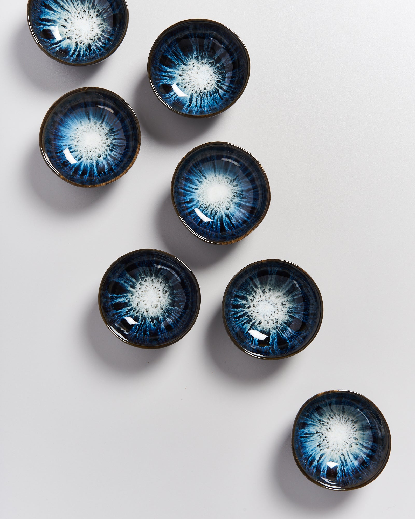 Sieben blaue, kunstvoll glasierte Keramikschalen mit strahlenförmigem Muster auf grauem Untergrund.