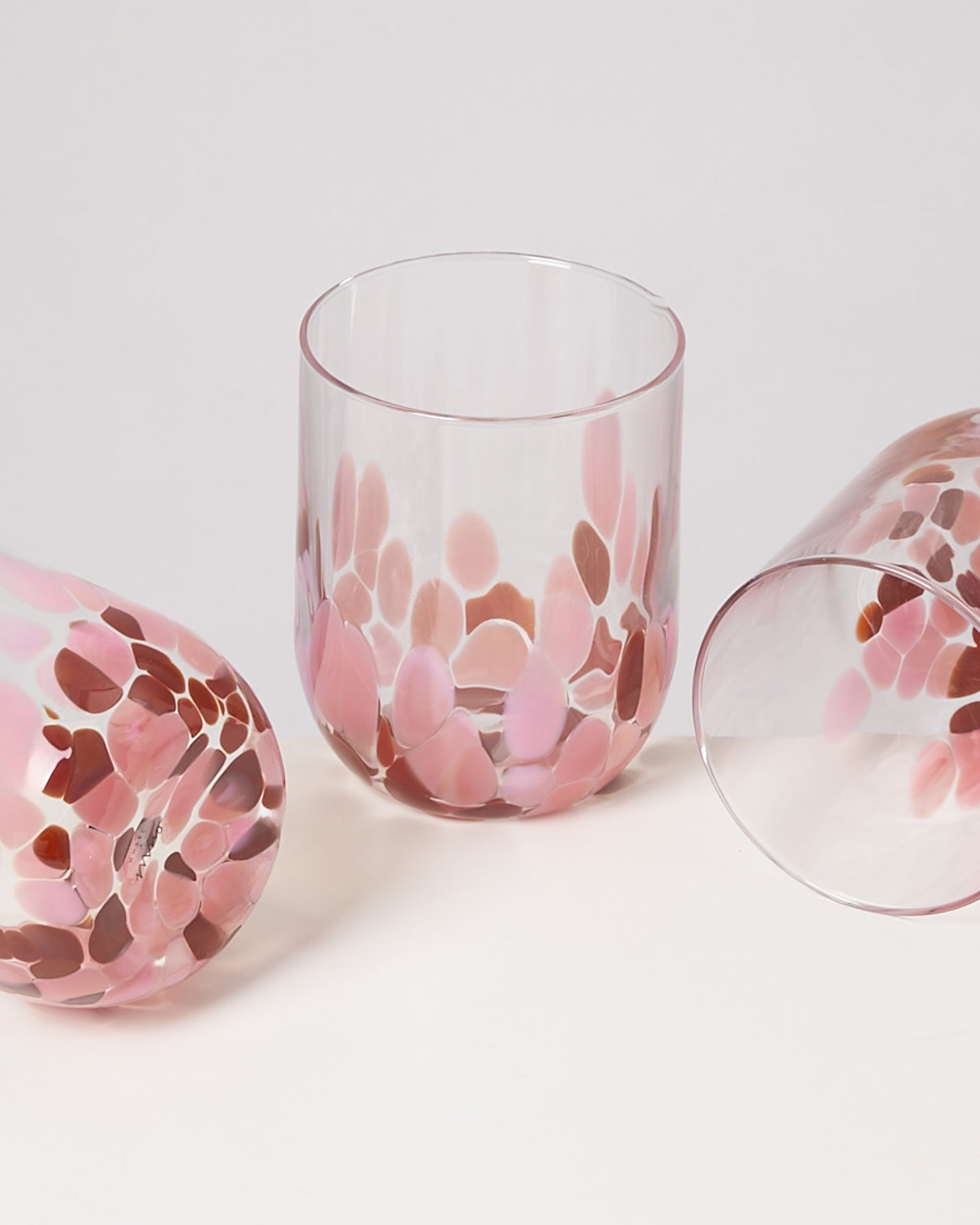 Drei Gläser mit rosa und braunem Tupfenmuster auf einer weißen Oberfläche, eines steht, zwei liegen.