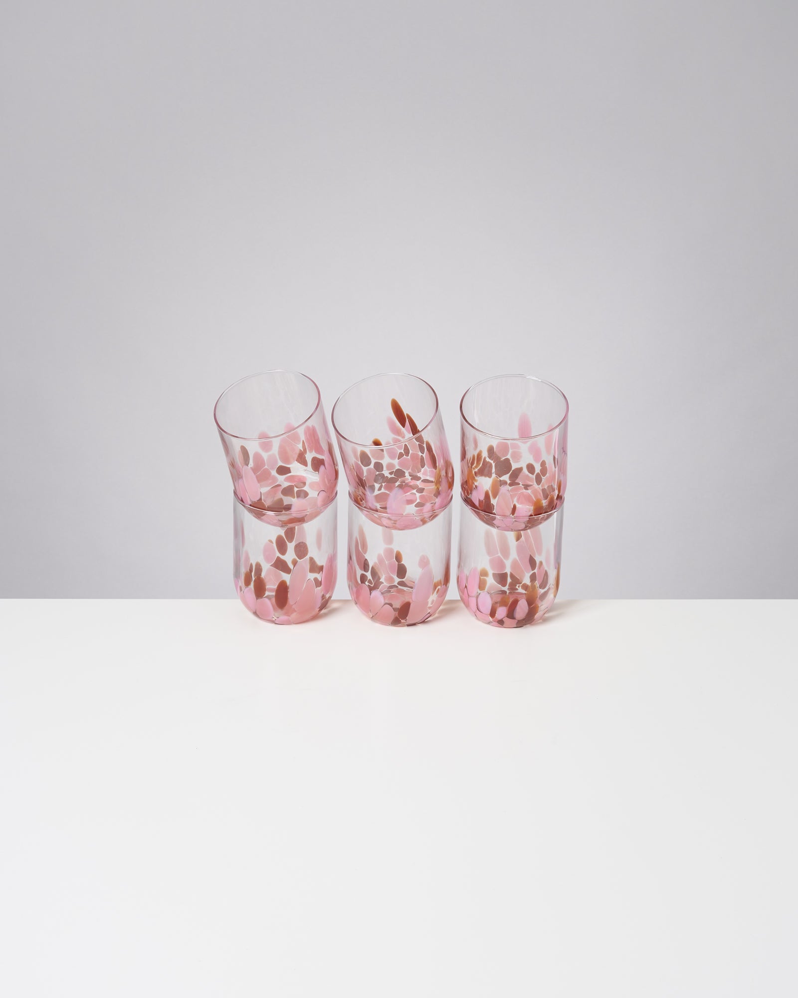 Drei Glasbecher mit rosa und braunen Mustern stehen auf einem weißen Tisch vor einem grauen Hintergrund.