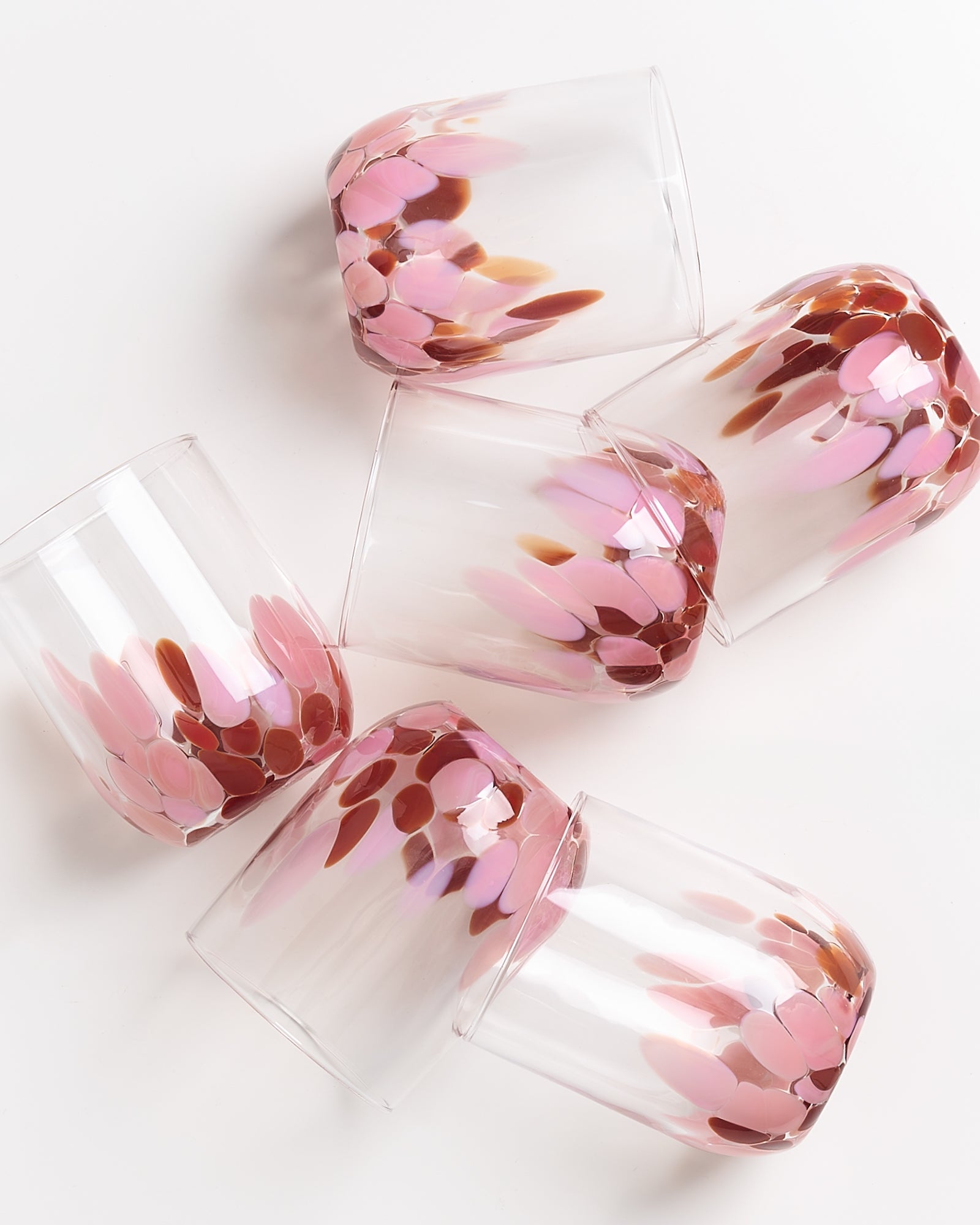 Fünf transparente Gläser mit rosa und braunen Farbmustern liegen schräg auf einer weißen Oberfläche.
