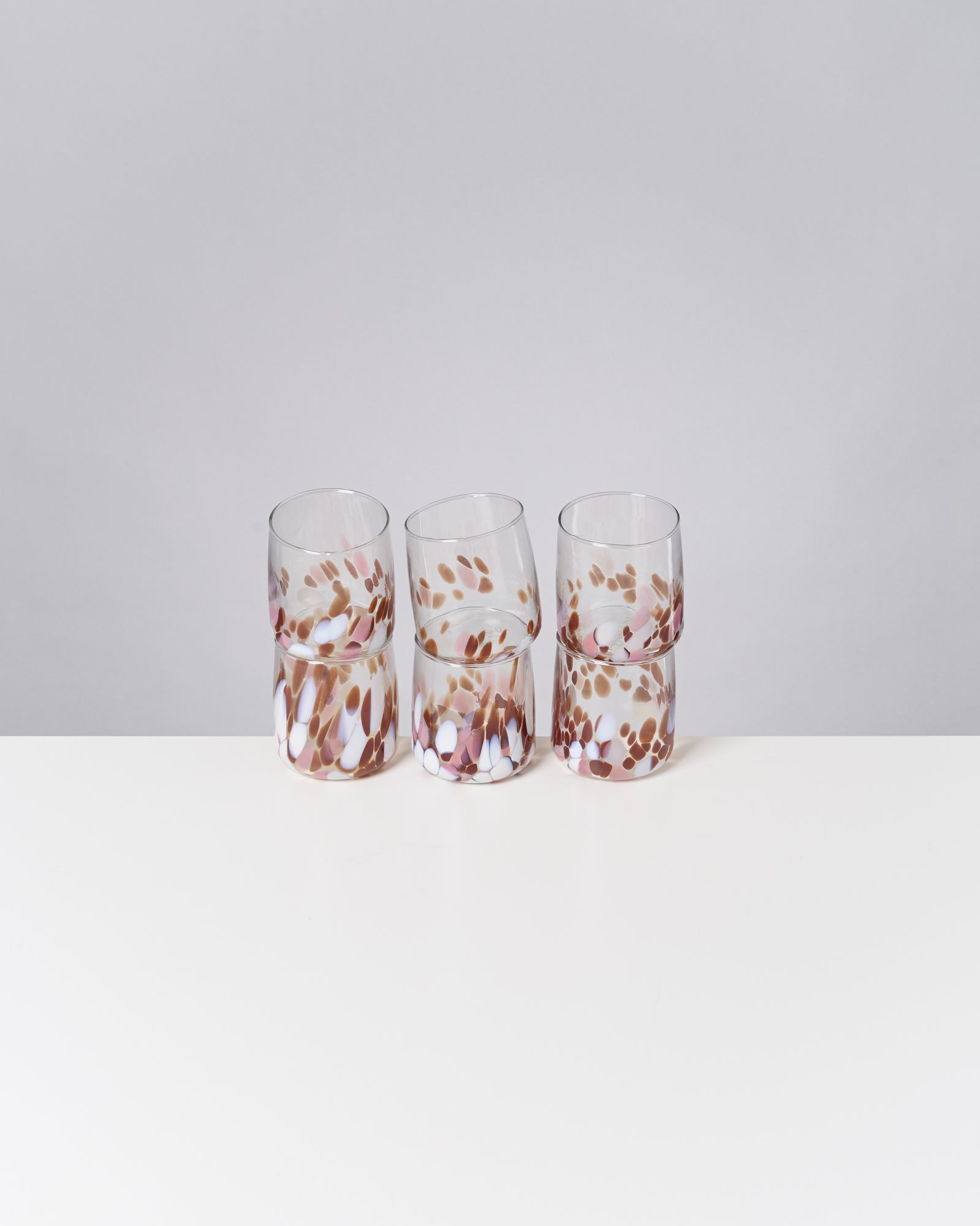 Sechs bunte Gläser mit fleckigem Muster stehen gestapelt auf einem weißen Tisch vor einem grauen Hintergrund.