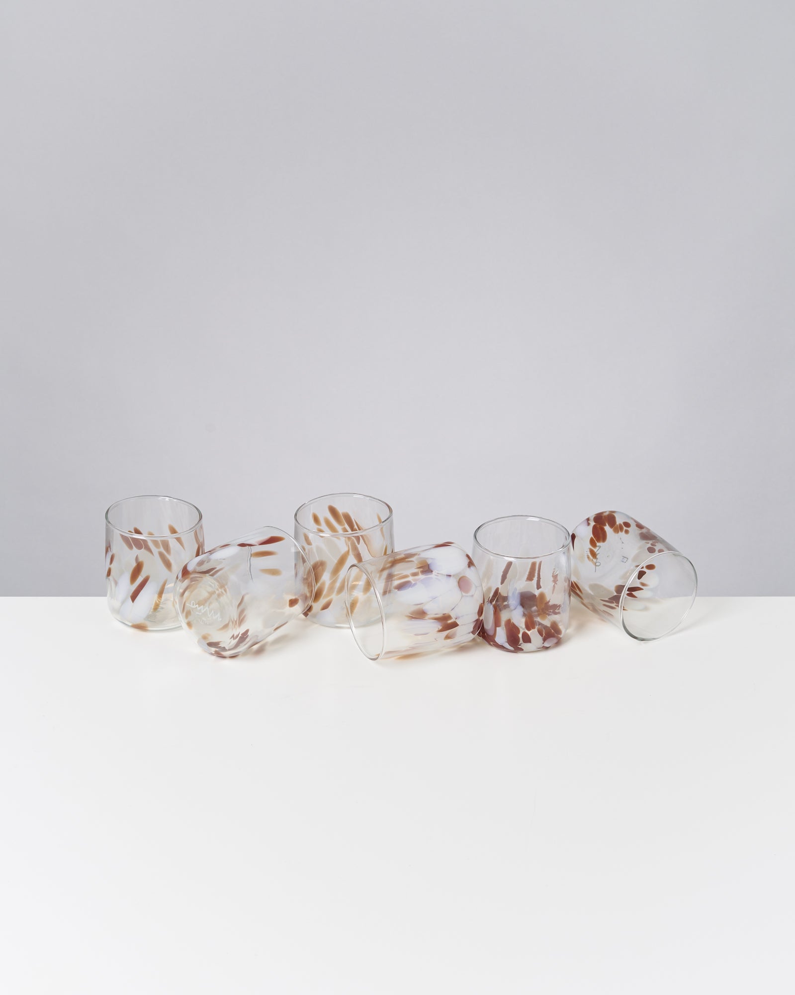 Sechs bunt gemusterte Gläser mit braunen und weißen Akzenten liegen auf einer weißen Oberfläche.