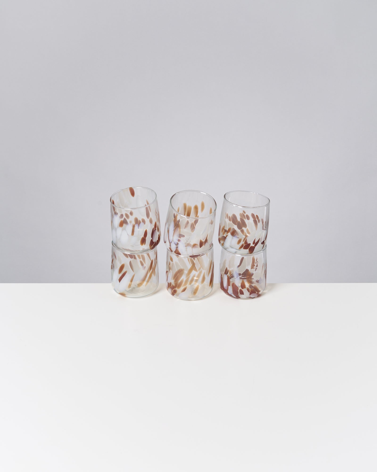 Sechs bunt gemusterte Gläser stehen in zwei Reihen auf einem weißen Tisch vor grauem Hintergrund.