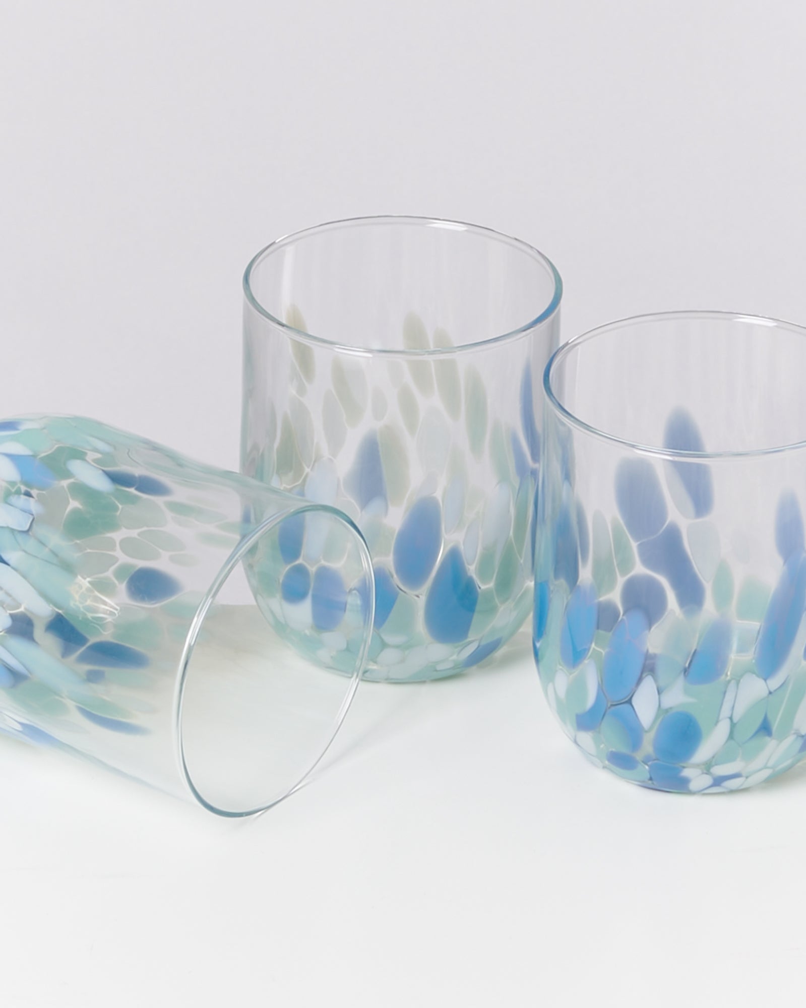 Drei Glasbecher mit blauen und grünen Flecken, zwei stehen aufrecht, einer liegt auf der Seite.