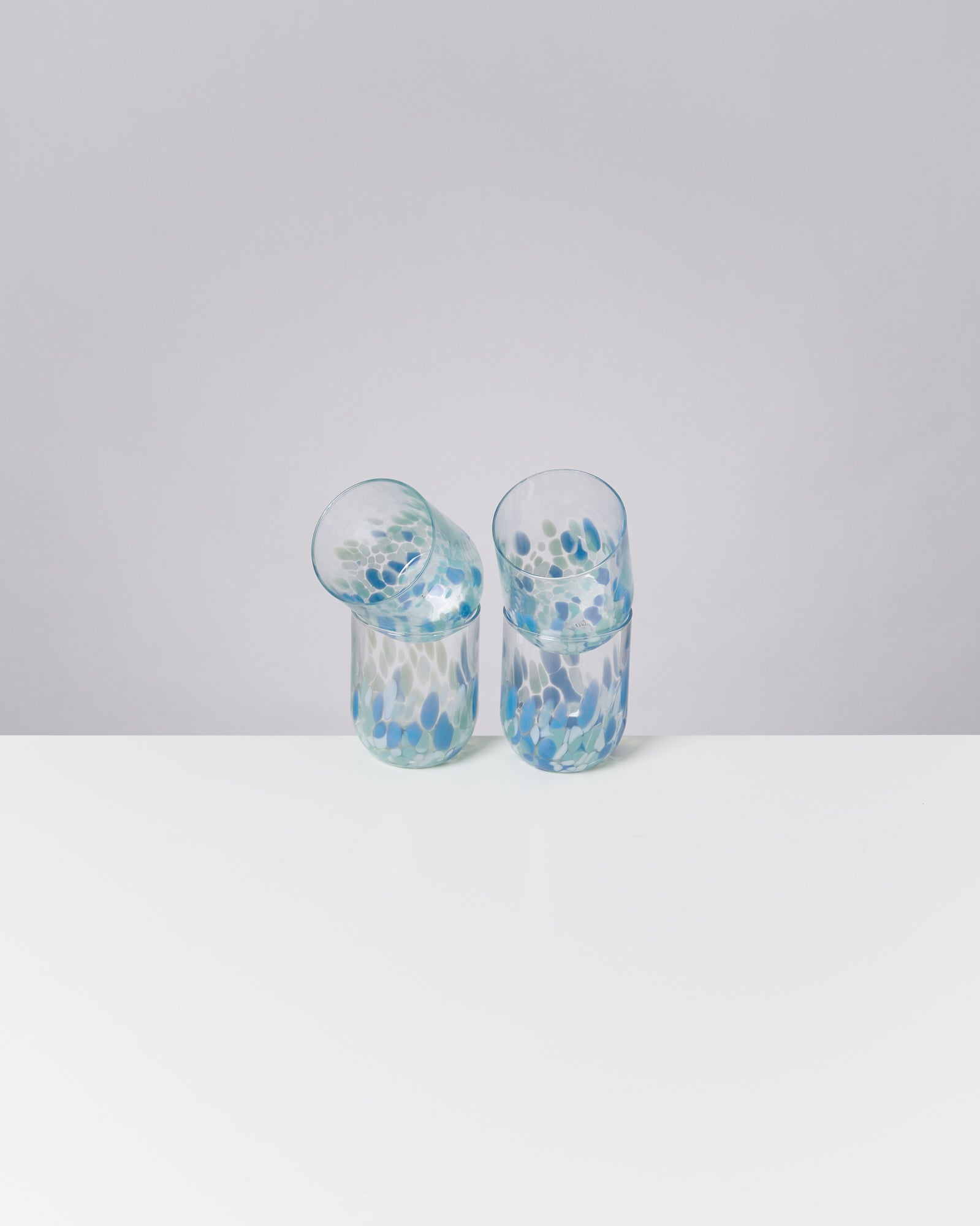 Zwei bunte Gläser mit blauen und grünen Mustern stehen auf einem weißen Tisch vor einem grauen Hintergrund.