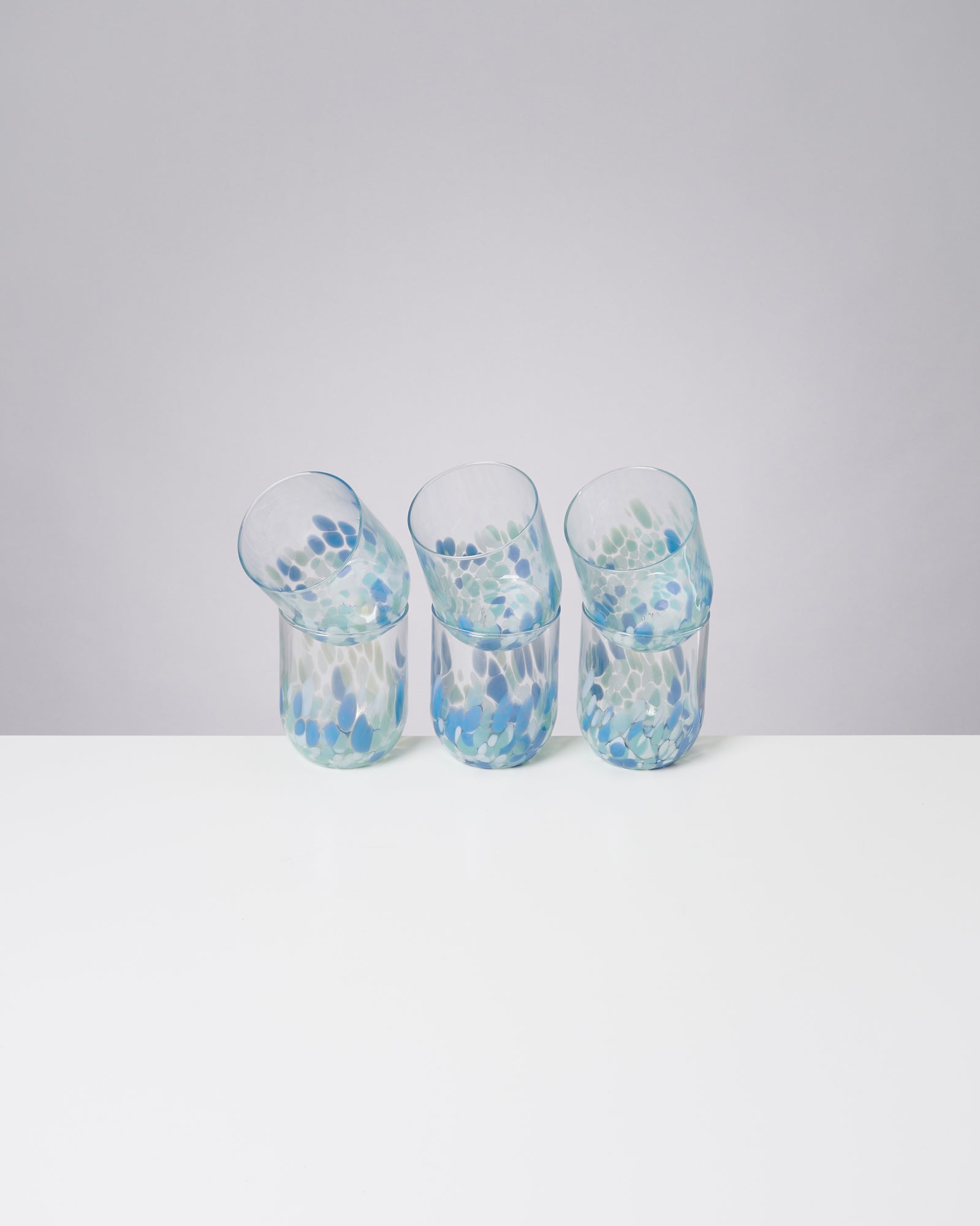 Drei bunte, gebogene Trinkgläser mit blauen und grünen Mustern stehen auf einem weißen Tisch vor grauem Hintergrund.