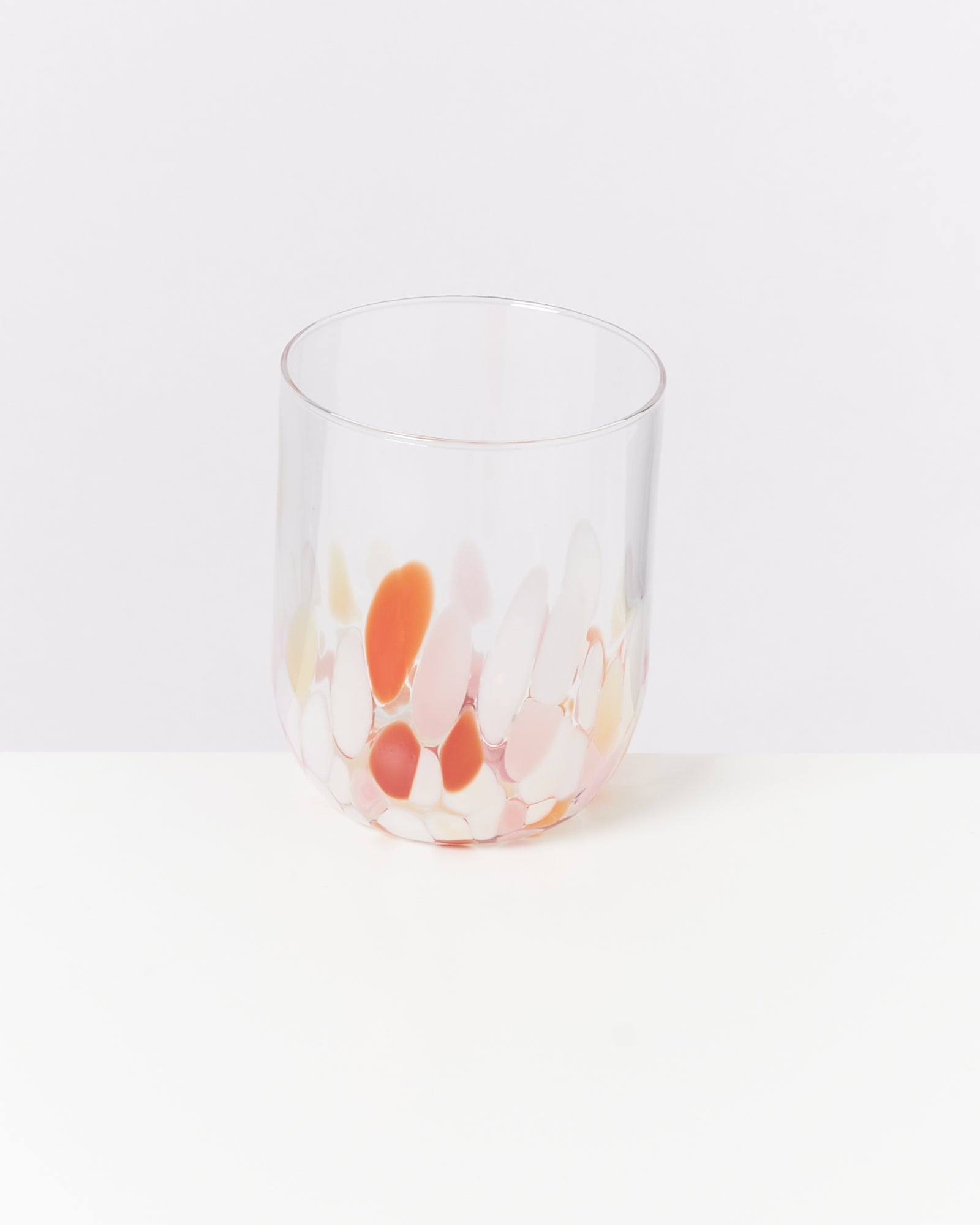 Ein transparentes Glas mit bunten, ovalen Farbklecksen in Rosa, Orange und Weiß auf weißem Hintergrund.