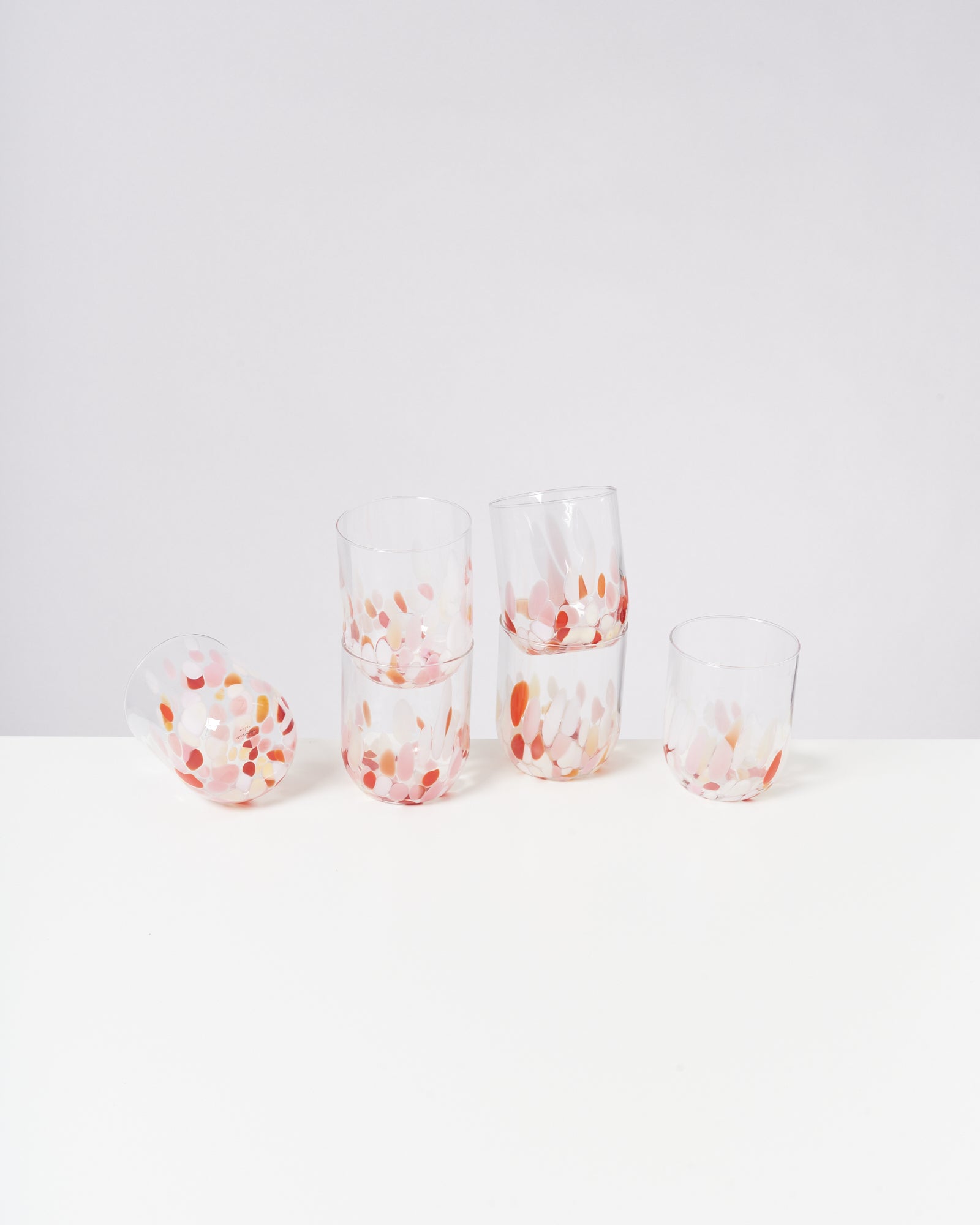 Fünf Glasbecher mit buntem Tupfenmuster stehen und liegen auf einem weißen Tisch vor hellem Hintergrund.