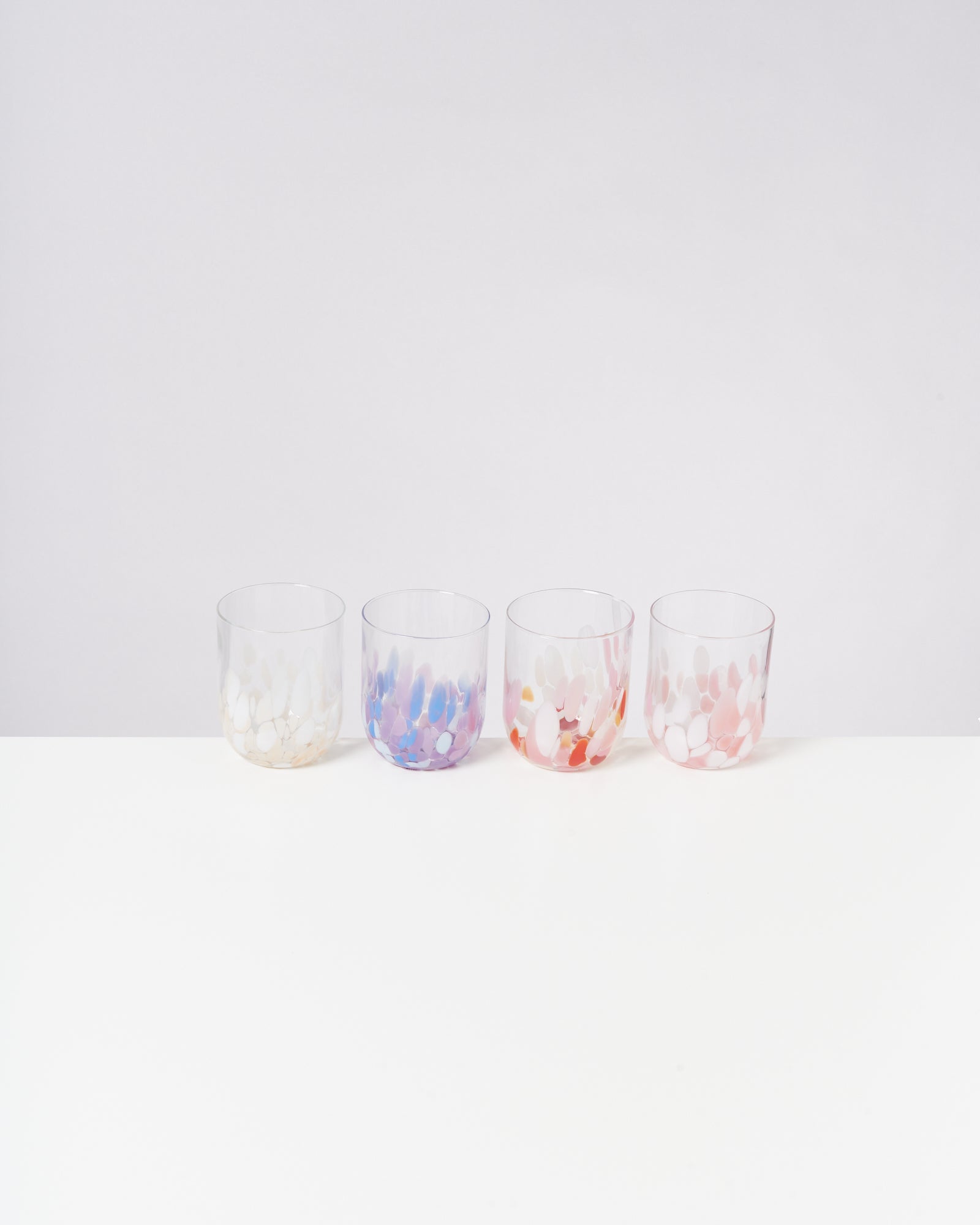 Vier bunte Trinkgläser mit getupften Mustern stehen auf einem weißen Tisch vor einem hellen Hintergrund.