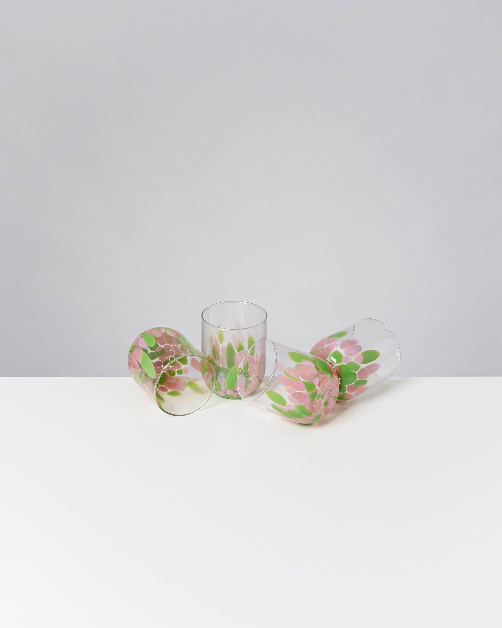 Drei gläserne Becher mit rosa und grünen Mustern liegen auf einem weißen Tisch vor grauem Hintergrund.