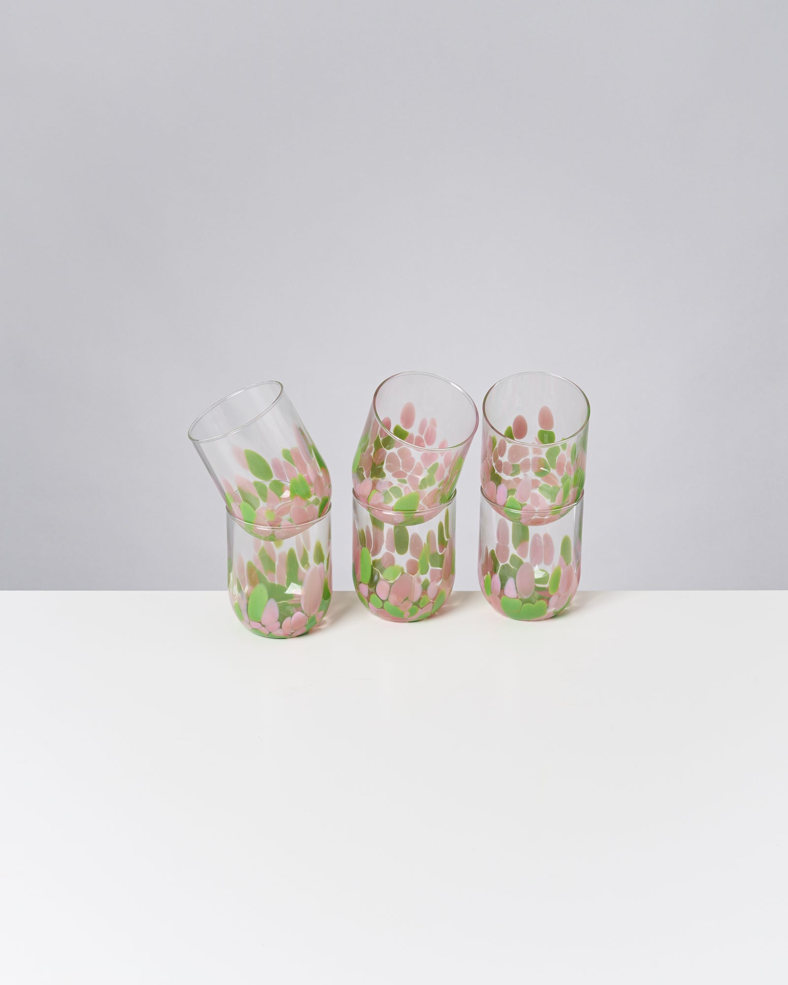 Drei bunt bemalte Glasbecher mit rosa und grünen Flecken stehen auf einem weißen Tisch vor einem grauen Hintergrund.