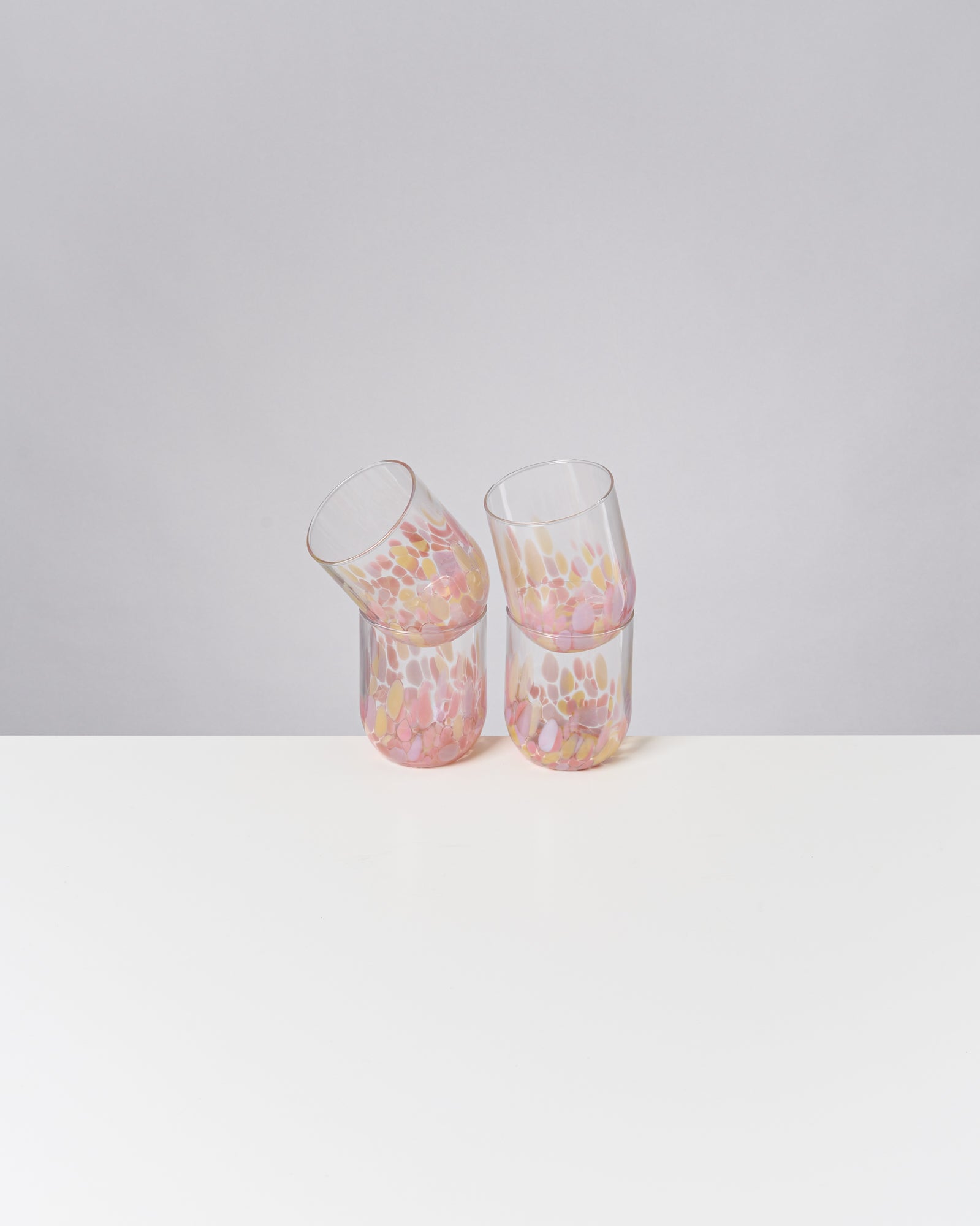 Vier bunte Gläser mit pastellfarbenem Muster stehen auf einem weißen Tisch vor grauem Hintergrund.