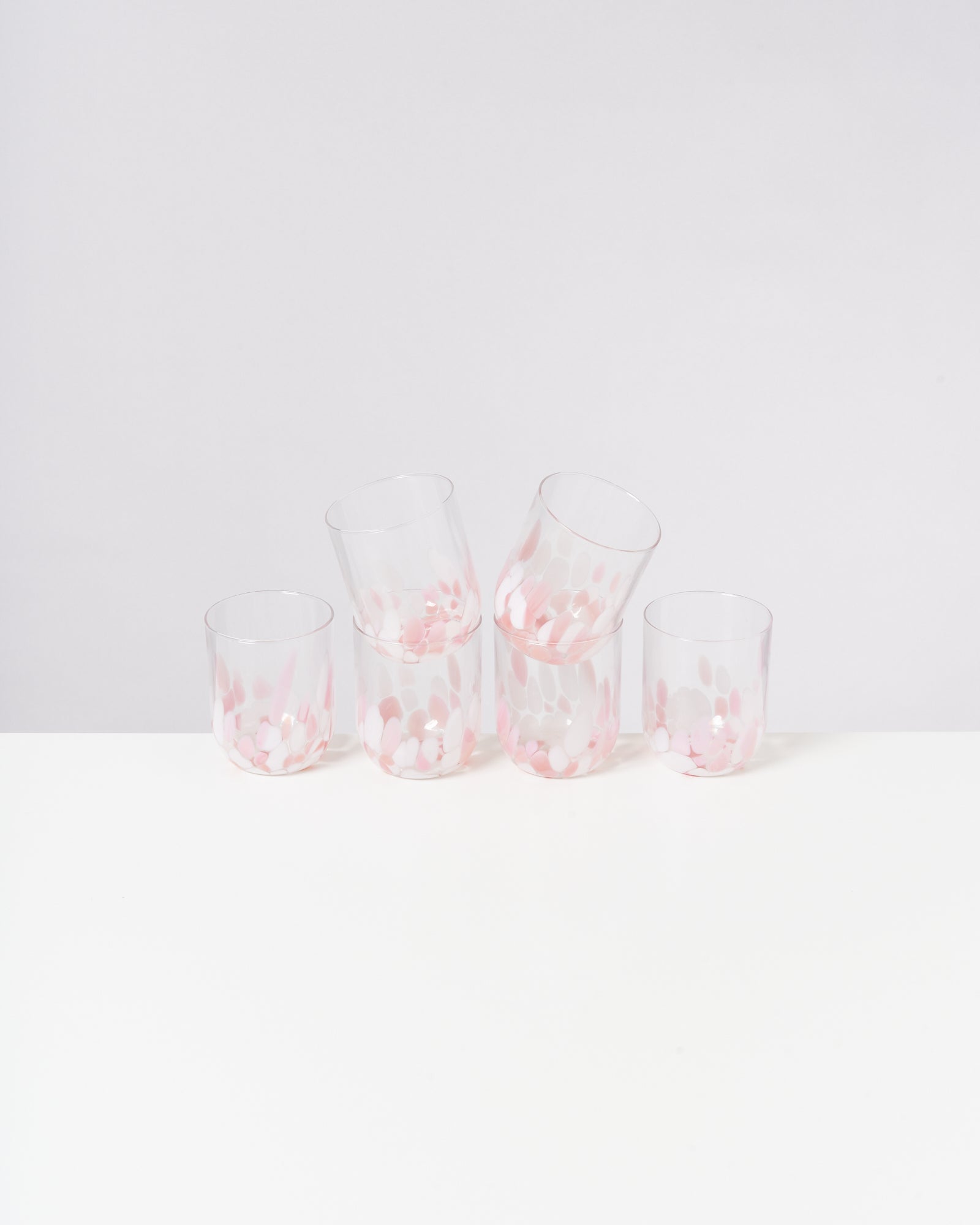 Fünf transparente Gläser mit rosa-weißen Mustern stehen auf einem weißen Tisch vor einer grauen Wand.