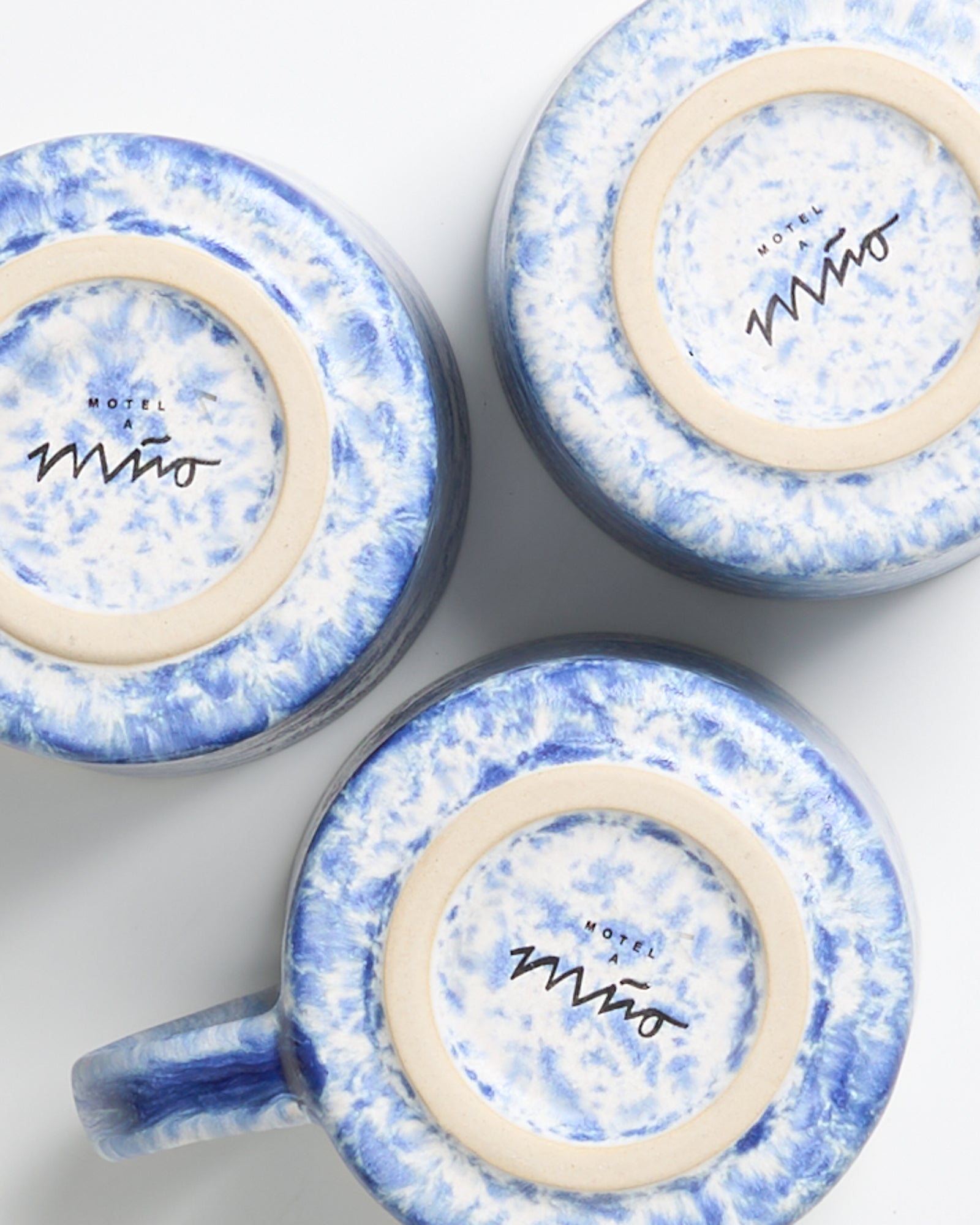 Drei blau-weiß marmorierte Keramiktassen mit "Motel a Mio" auf der Unterseite, von oben fotografiert.