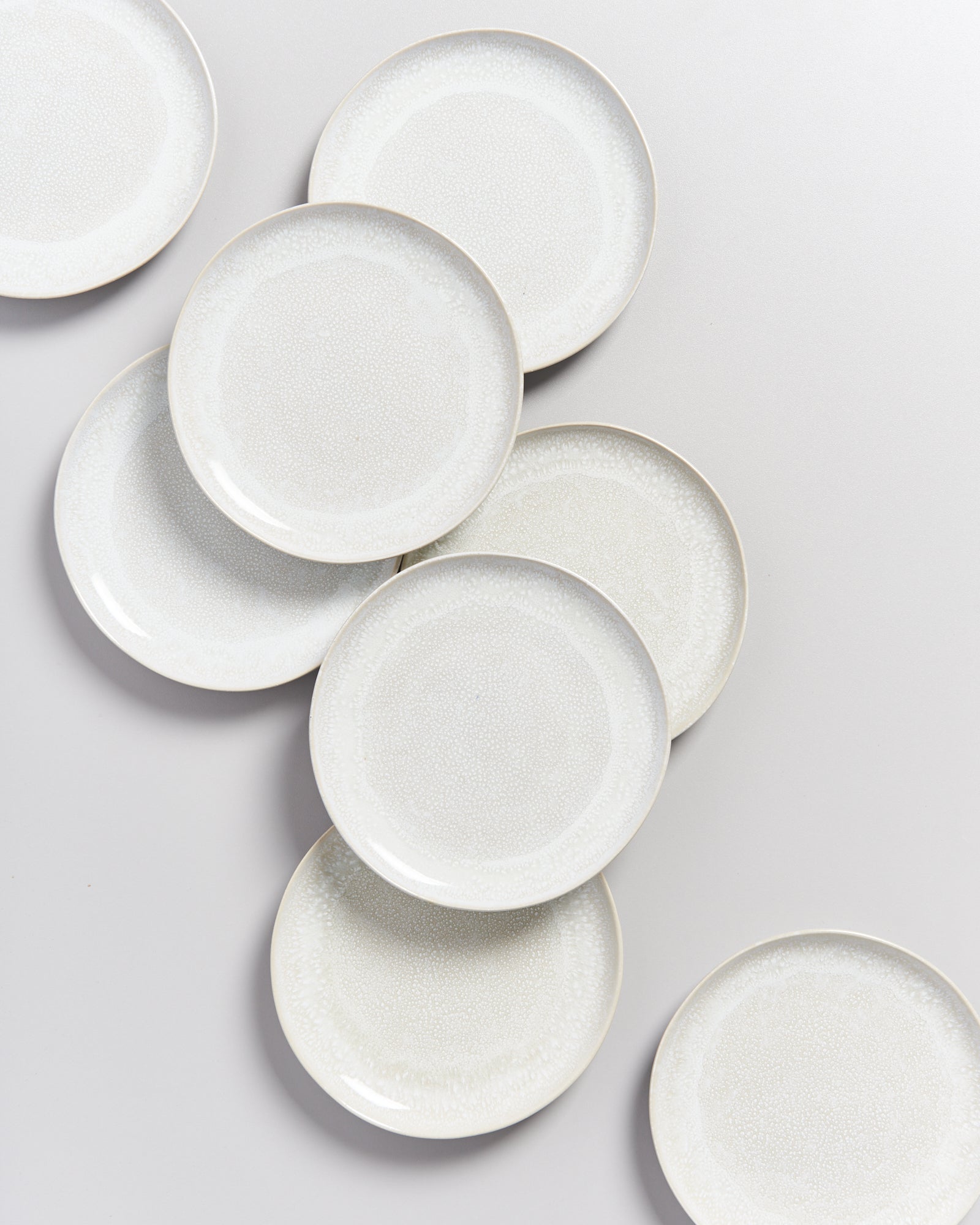Acht weiße Teller mit leichtem Muster sind unregelmäßig auf einer hellen Oberfläche angeordnet.