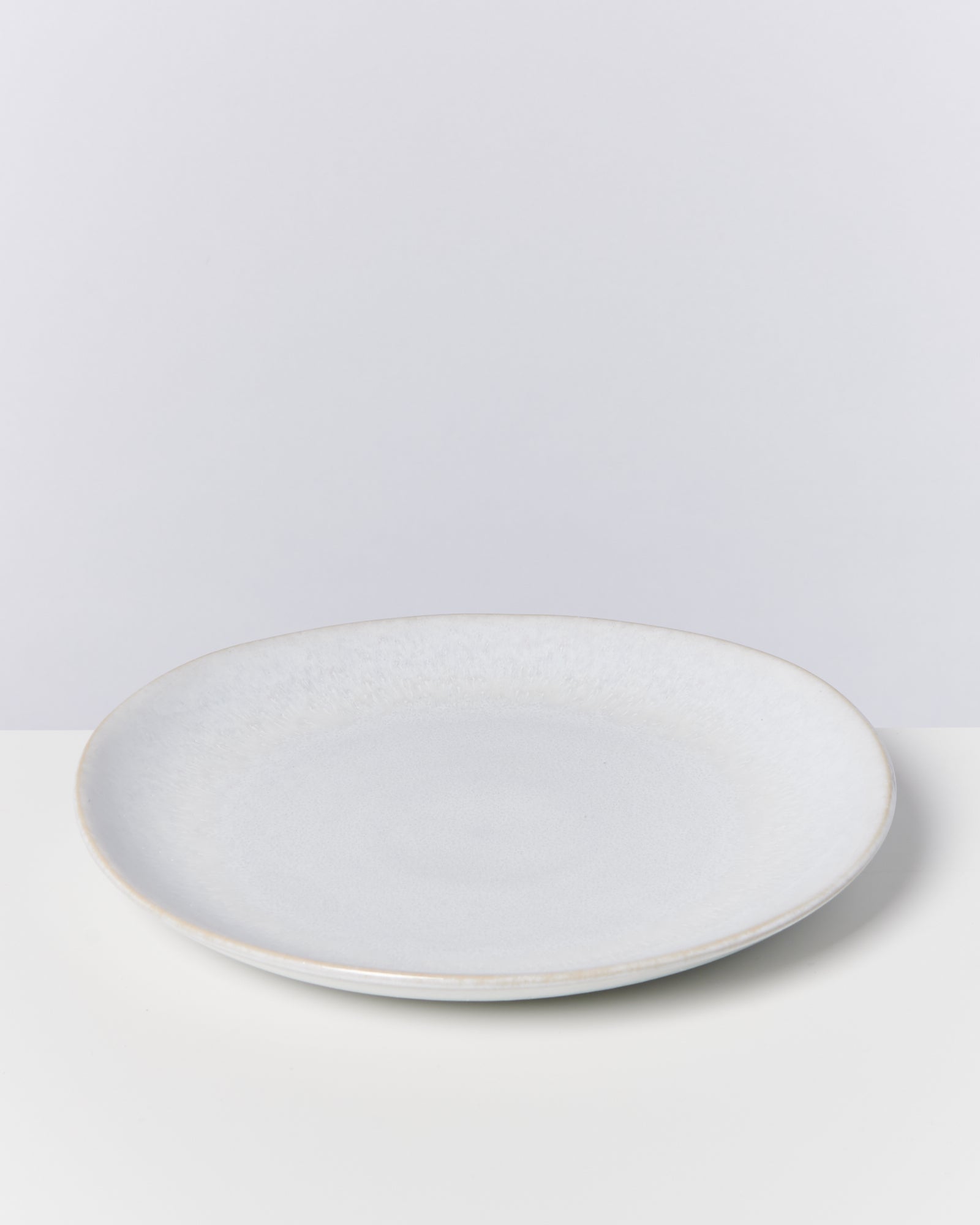 Ein heller, flacher Keramikteller steht auf einem weißen Untergrund vor einer grauen Wand.