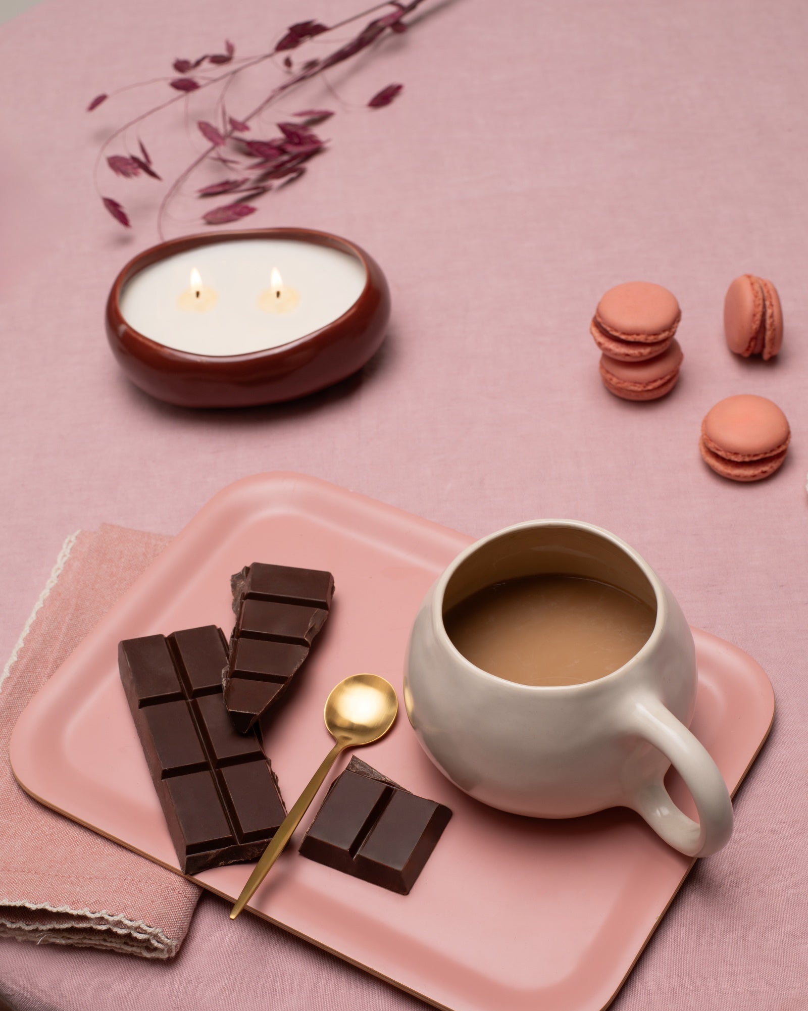 Ein Tisch mit Kerze, Schokolade, einer Tasse Kaffee, einem goldenen Löffel und rosafarbenen Macarons auf rosa Tischdecke.