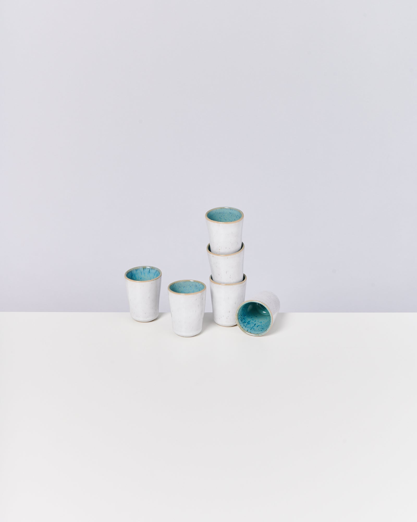 Sechs weiße Keramikbecher mit blauer Innenseite, fünf gestapelt und einer liegend, auf weißem Hintergrund.