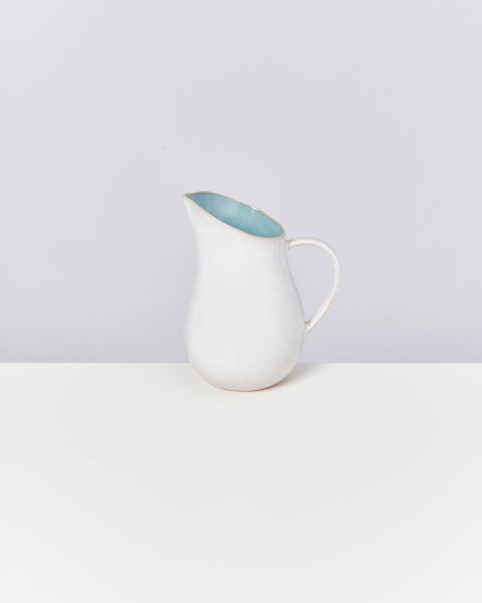 Weiße Keramikkanne mit hellblauem Inneren, auf einem weißen Tisch vor grauem Hintergrund.