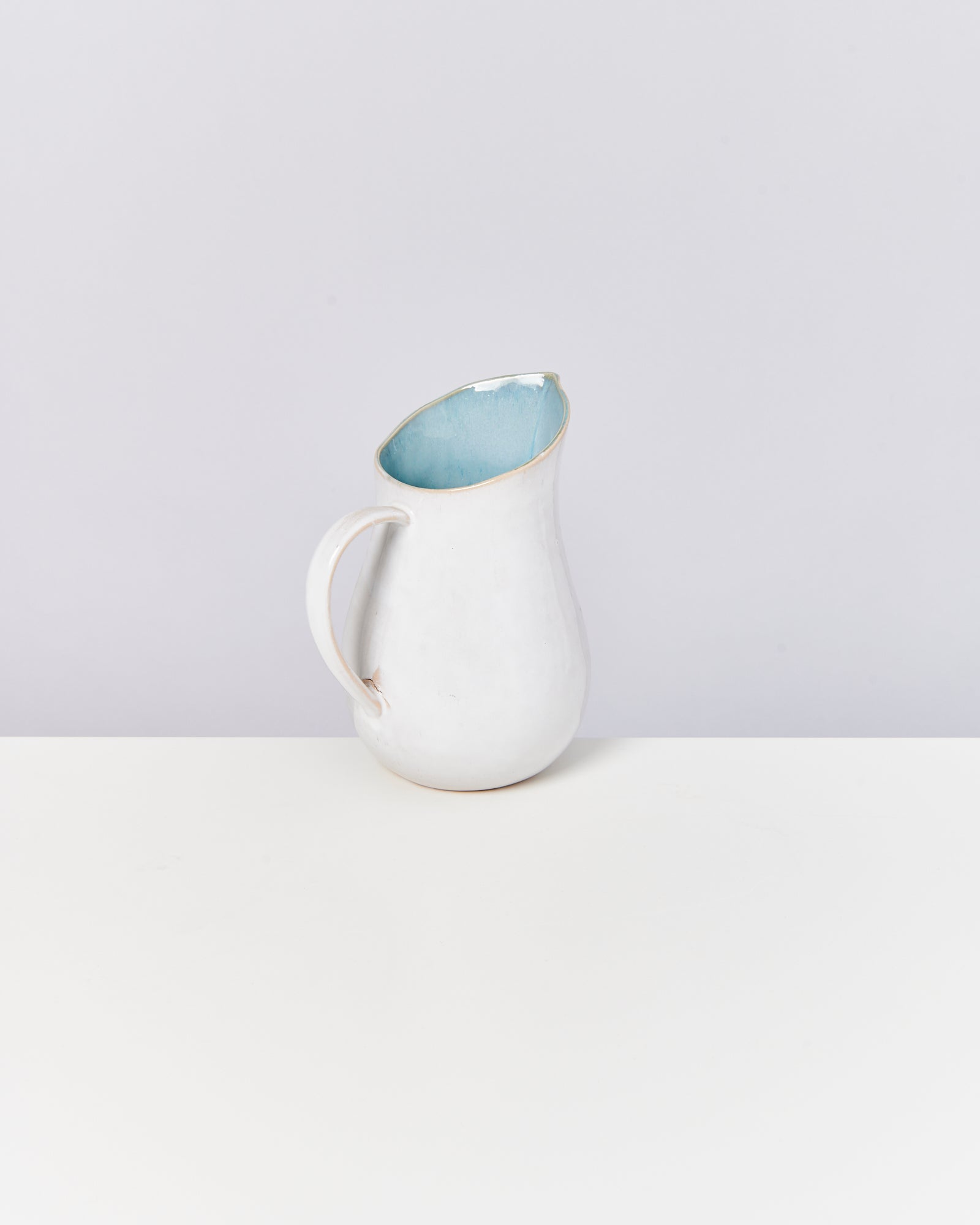 Ein weißer Krug mit hellem, blauem Inneren steht auf einem weißen Tisch vor einer grauen Wand.
