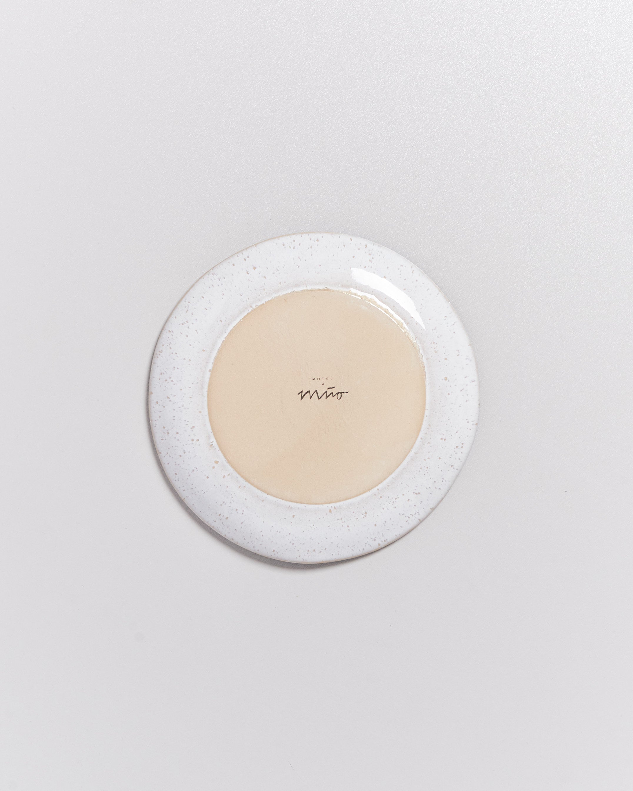 Eine umgedrehte, runde Keramikplatte mit beigem Zentrum und weißem, gesprenkeltem Rand auf grauem Hintergrund.