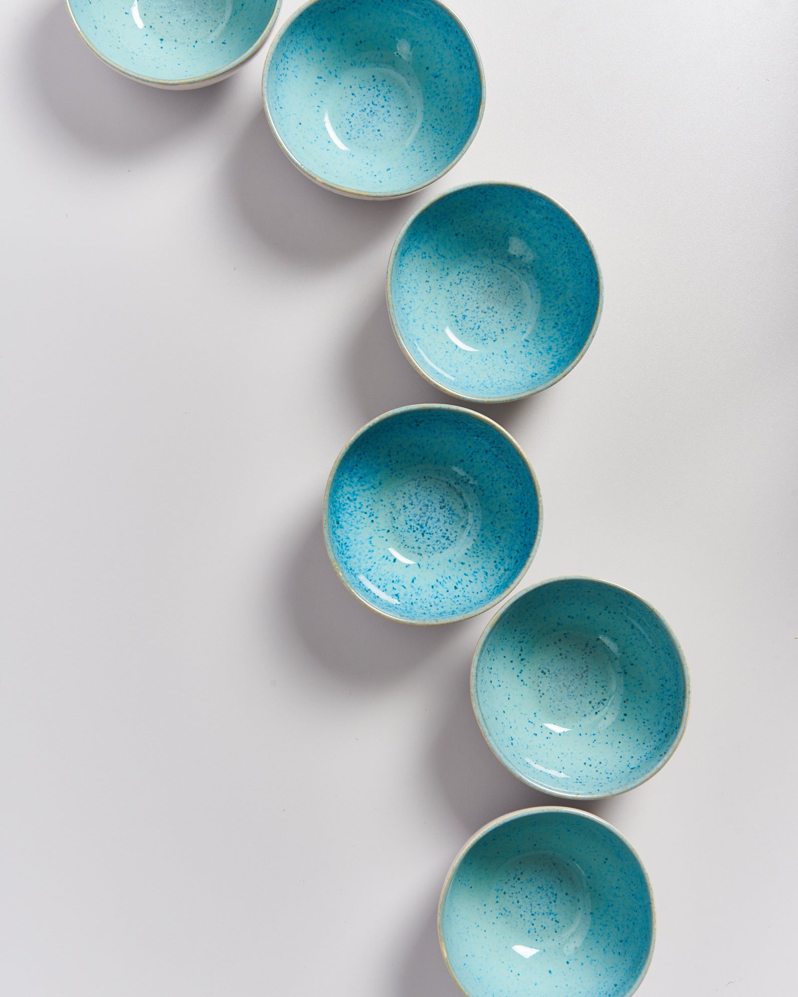 Sechs blaue Schalen mit gesprenkeltem Muster sind in einer geschlängelten Linie auf einer weißen Oberfläche angeordnet.