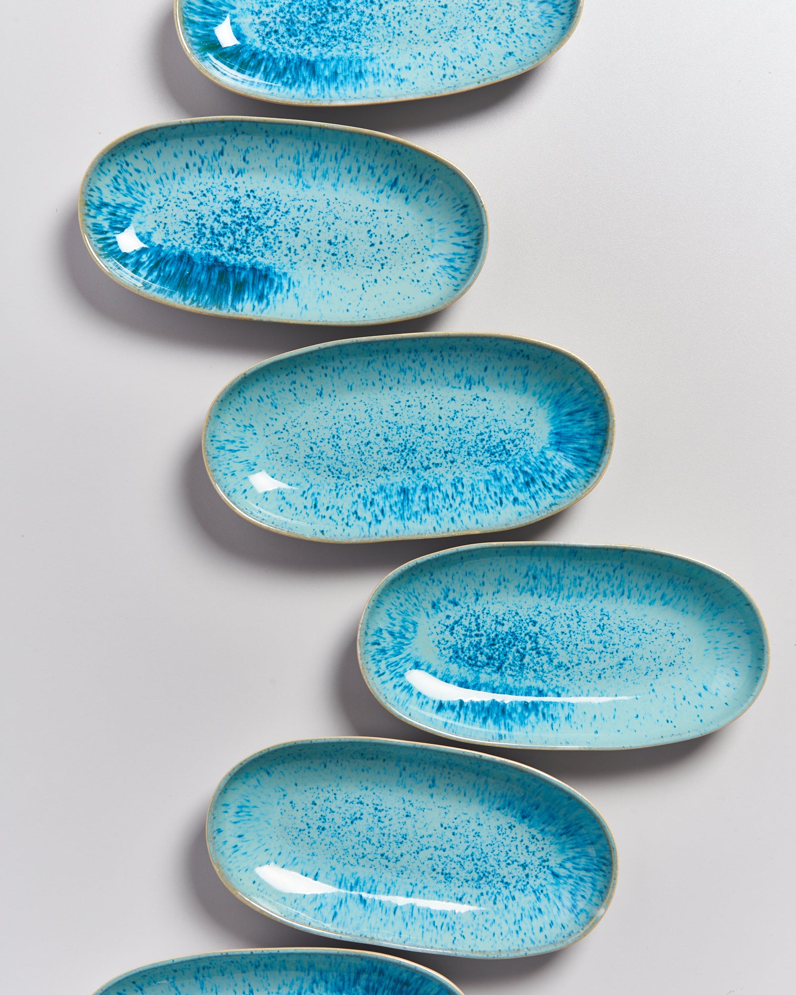 Sechs ovale, türkisblau gesprenkelte Teller sind schräg angeordnet auf einer weißen Oberfläche zu sehen.