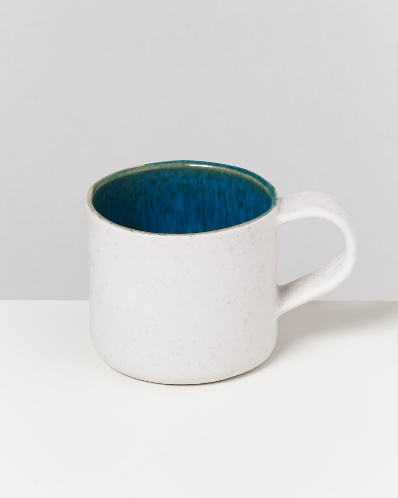 Weiße Keramiktasse mit blauem Innenraum auf weißer Oberfläche vor grauem Hintergrund.