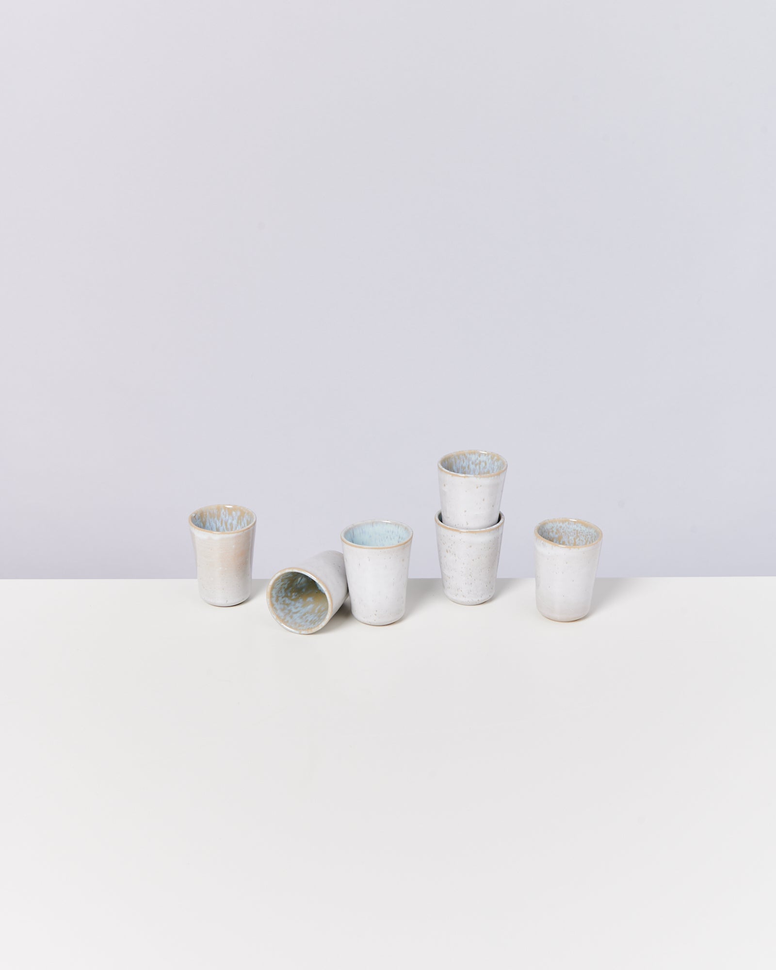 Sechs weiße Keramiktassen mit bläulichem Inneren, auf einem weißen Tisch vor grauem Hintergrund angeordnet.