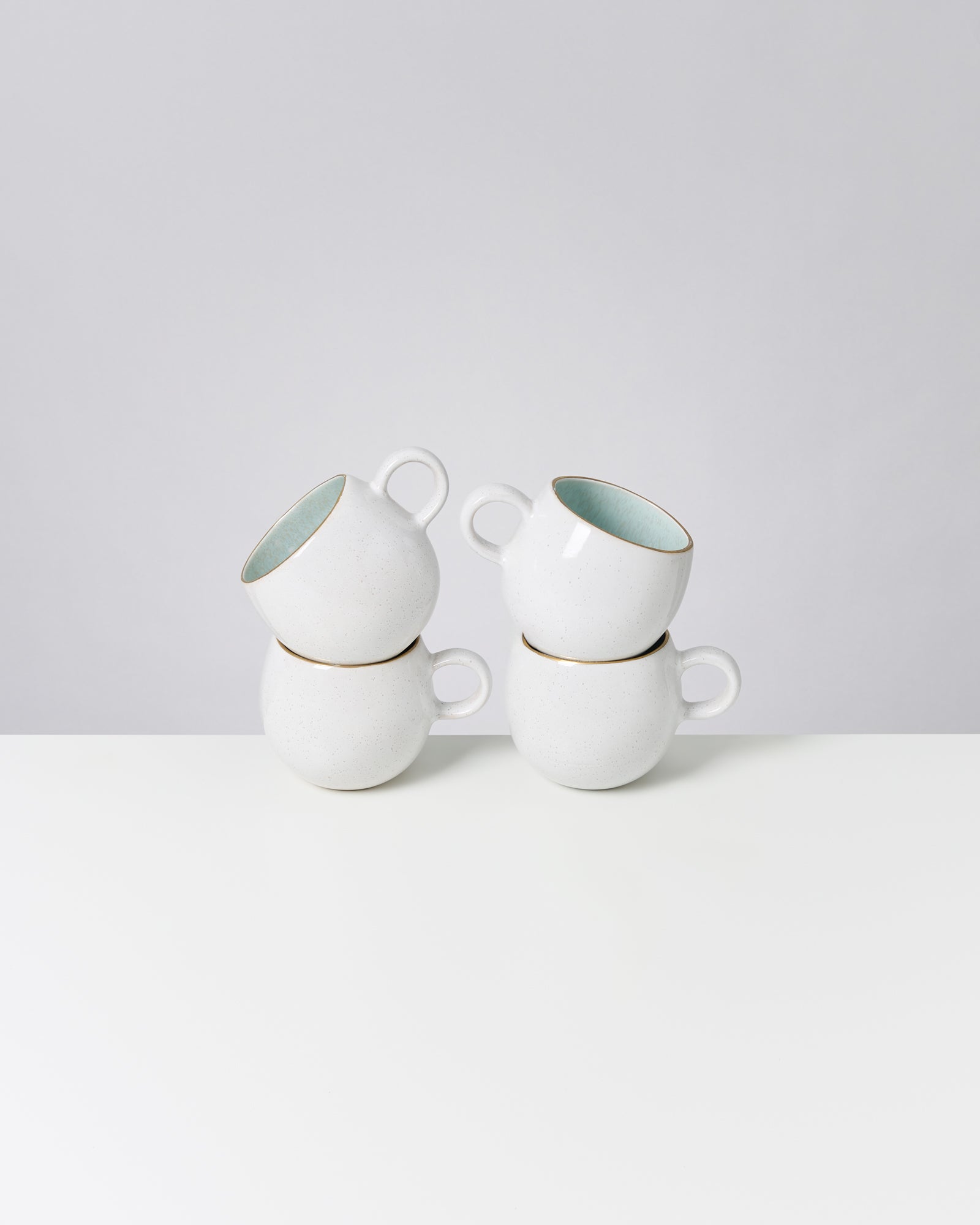 Vier gestapelte weiße Keramiktassen mit hellblauer Innenseite auf einem weißen Tisch.