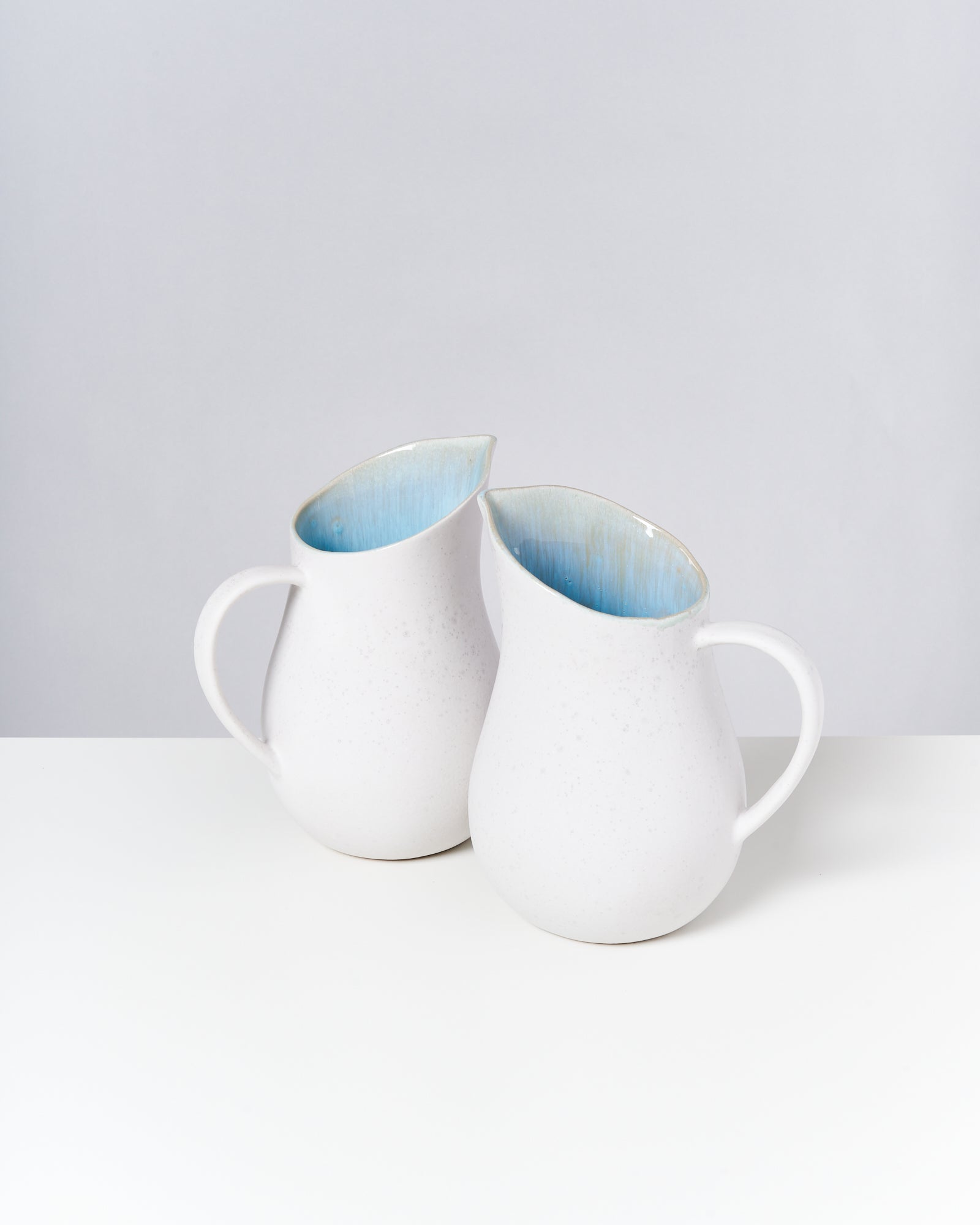 Zwei weiße Keramikkannen mit hellblauer Innenseite stehen auf einem weißen Tisch vor einem grauen Hintergrund.