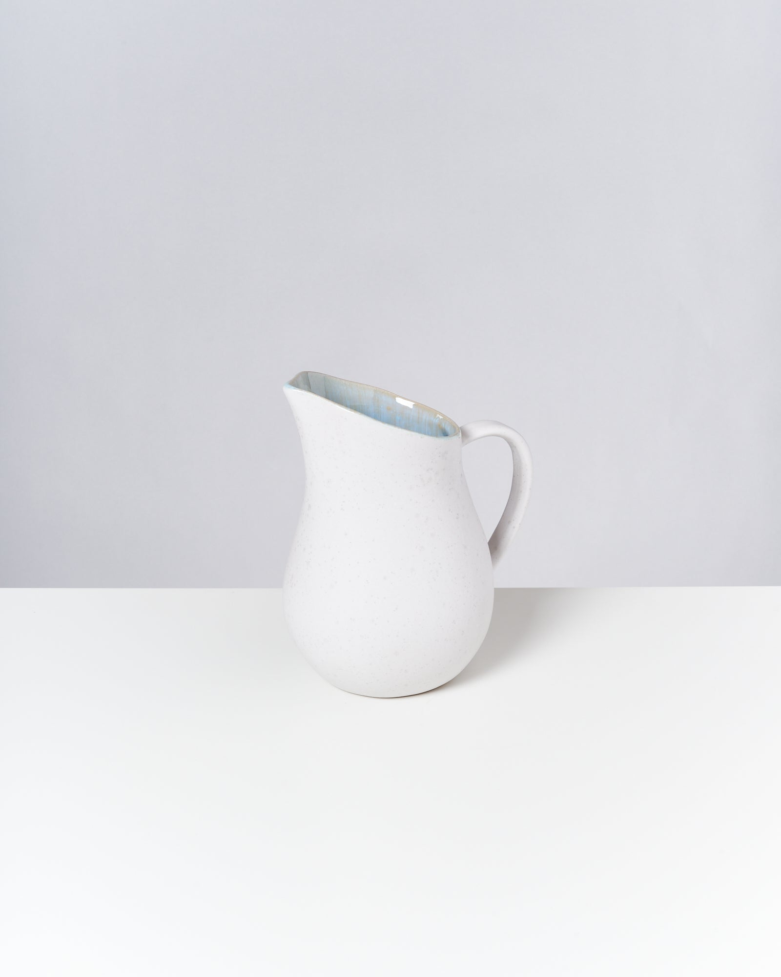 Ein weißer Krug mit Henkel steht auf einem weißen Tisch vor einem grauen Hintergrund.