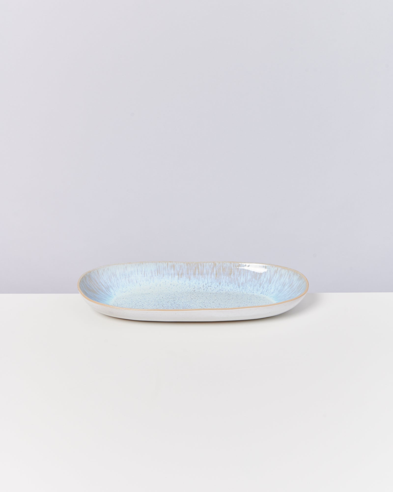 Ovaler, flacher Keramikteller mit blauer Glasur steht auf einer weißen Oberfläche vor neutralem Hintergrund.