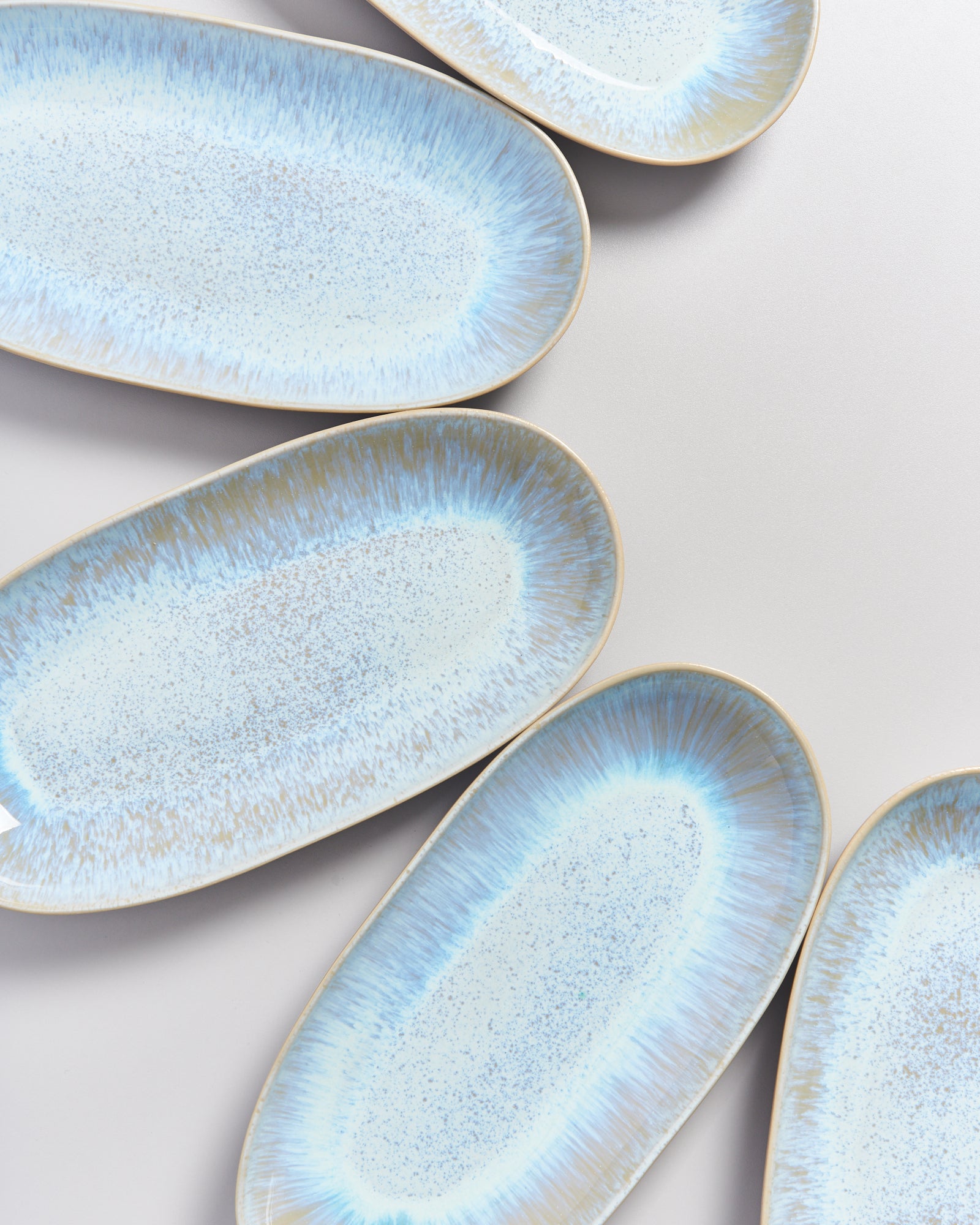 Fünf ovale, hellblaue Keramikteller mit bläulichem Randmuster liegen auf einer grauen Oberfläche.
