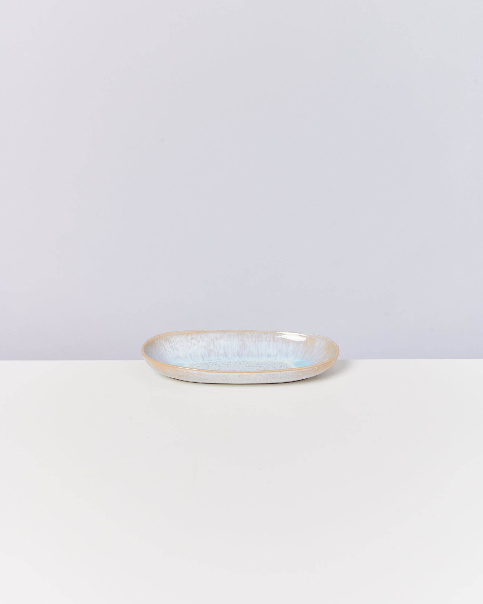 Ein ovaler Teller mit blauweißer Glasur steht auf einer weißen Oberfläche vor einem hellgrauen Hintergrund.