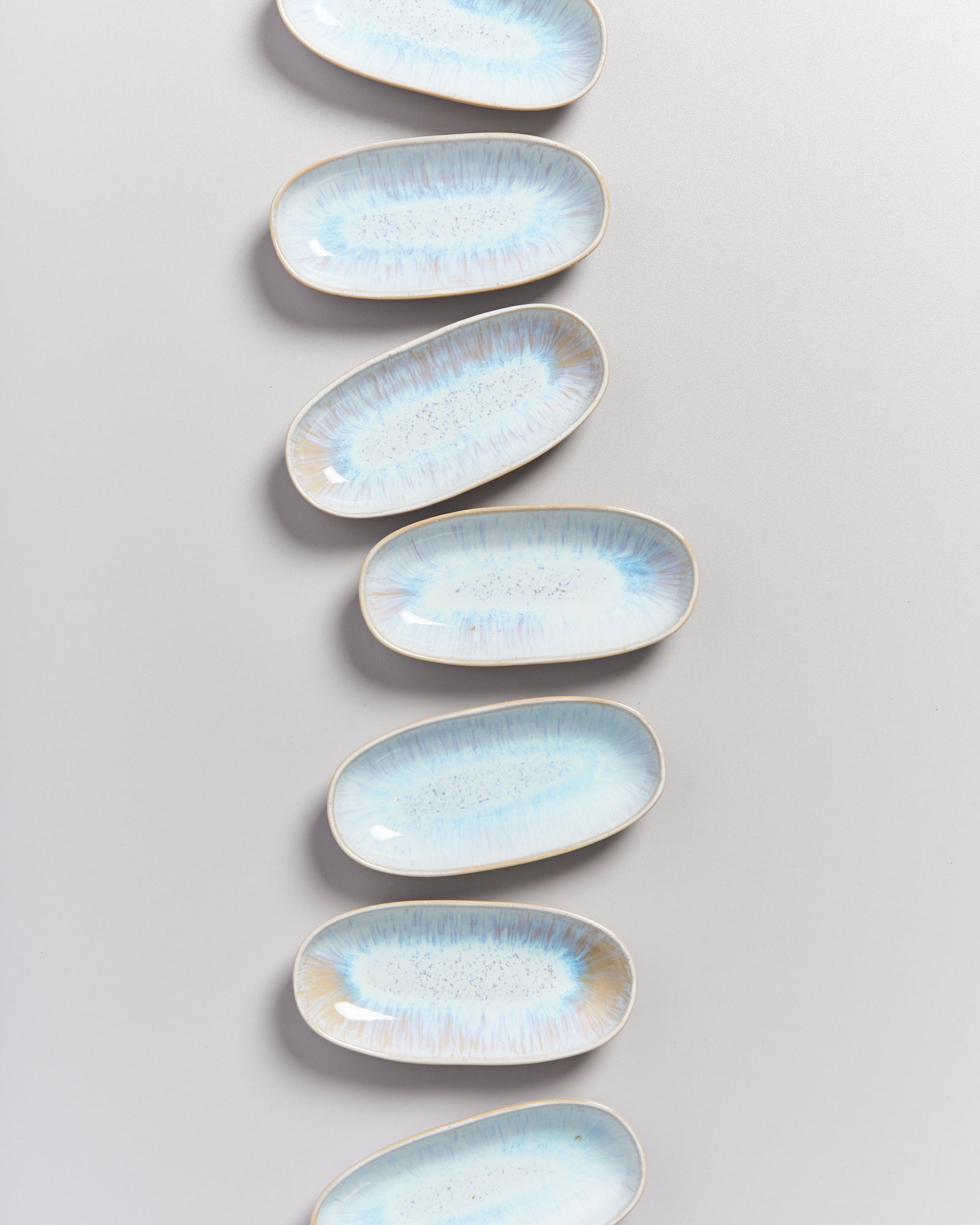 Acht ovale, blau-weiße Keramikteller sind in einer vertikalen Linie auf hellgrauem Hintergrund angeordnet.