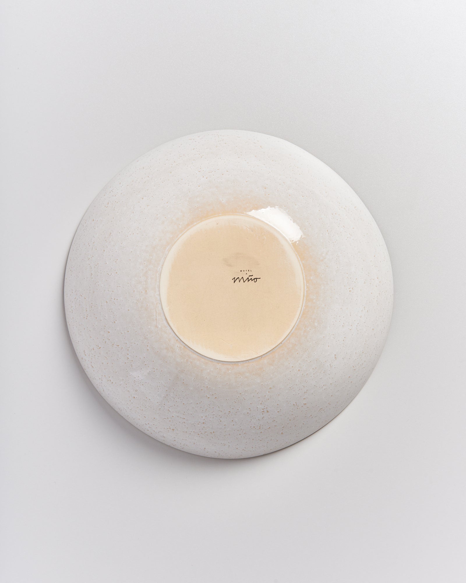 Eine weiße, runde Keramikschale mit Signatur in der Mitte, auf einem hellgrauen Hintergrund platziert.