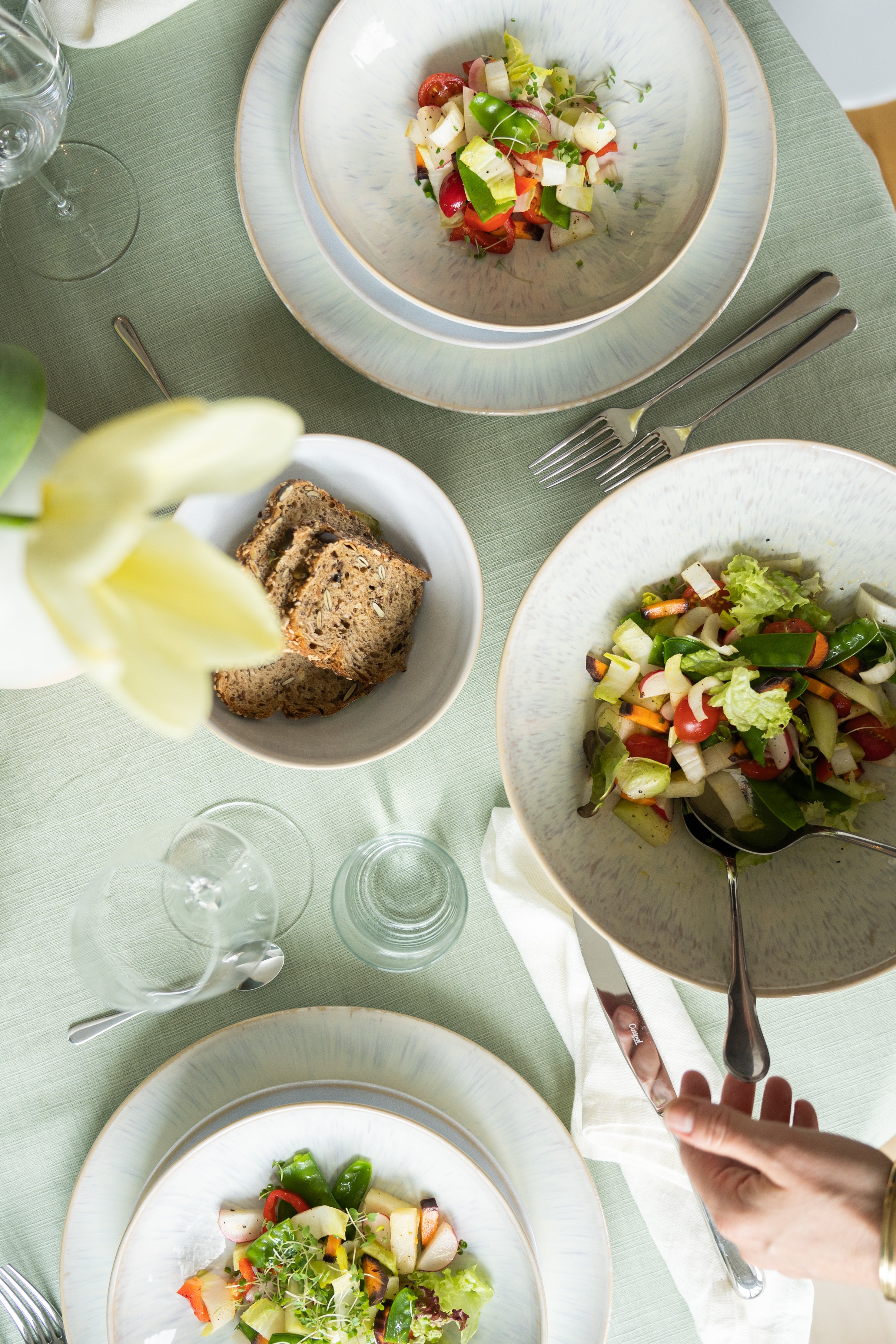 Mittagsgedeck: Drei Salatteller, Brotschale und Gläser auf grünem Tischtuch, Hand serviert.