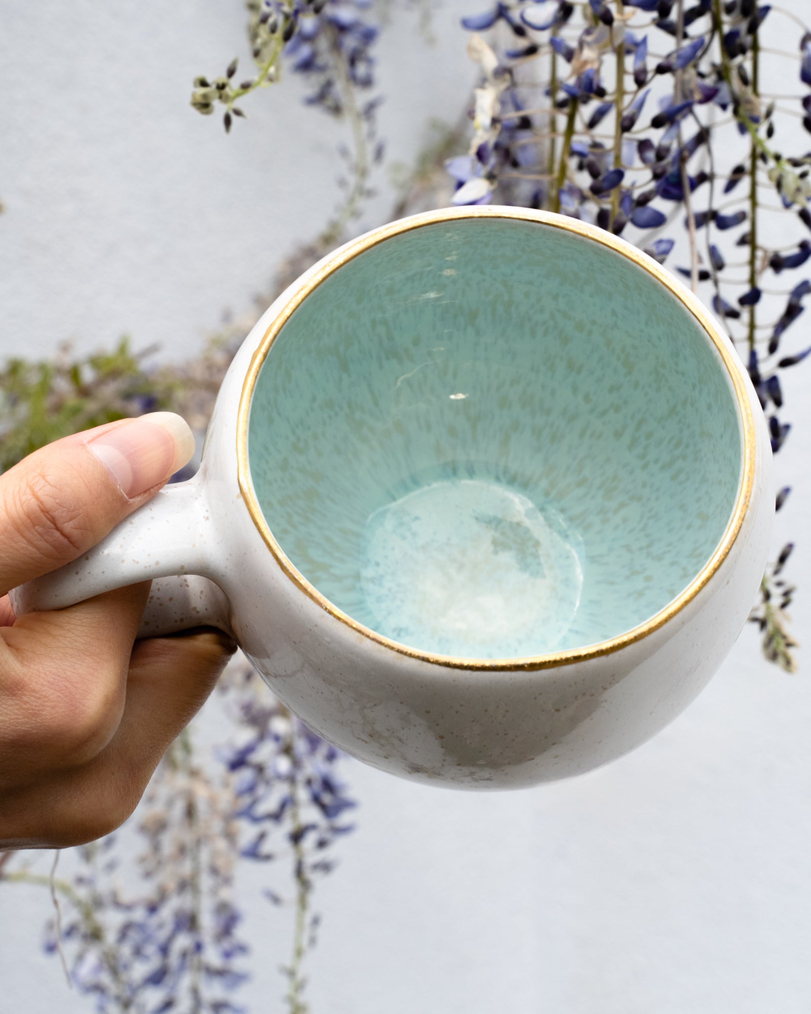 Hand hält eine Keramiktasse mit mintgrüner Innenseite vor blühenden lila Blumen im Hintergrund.