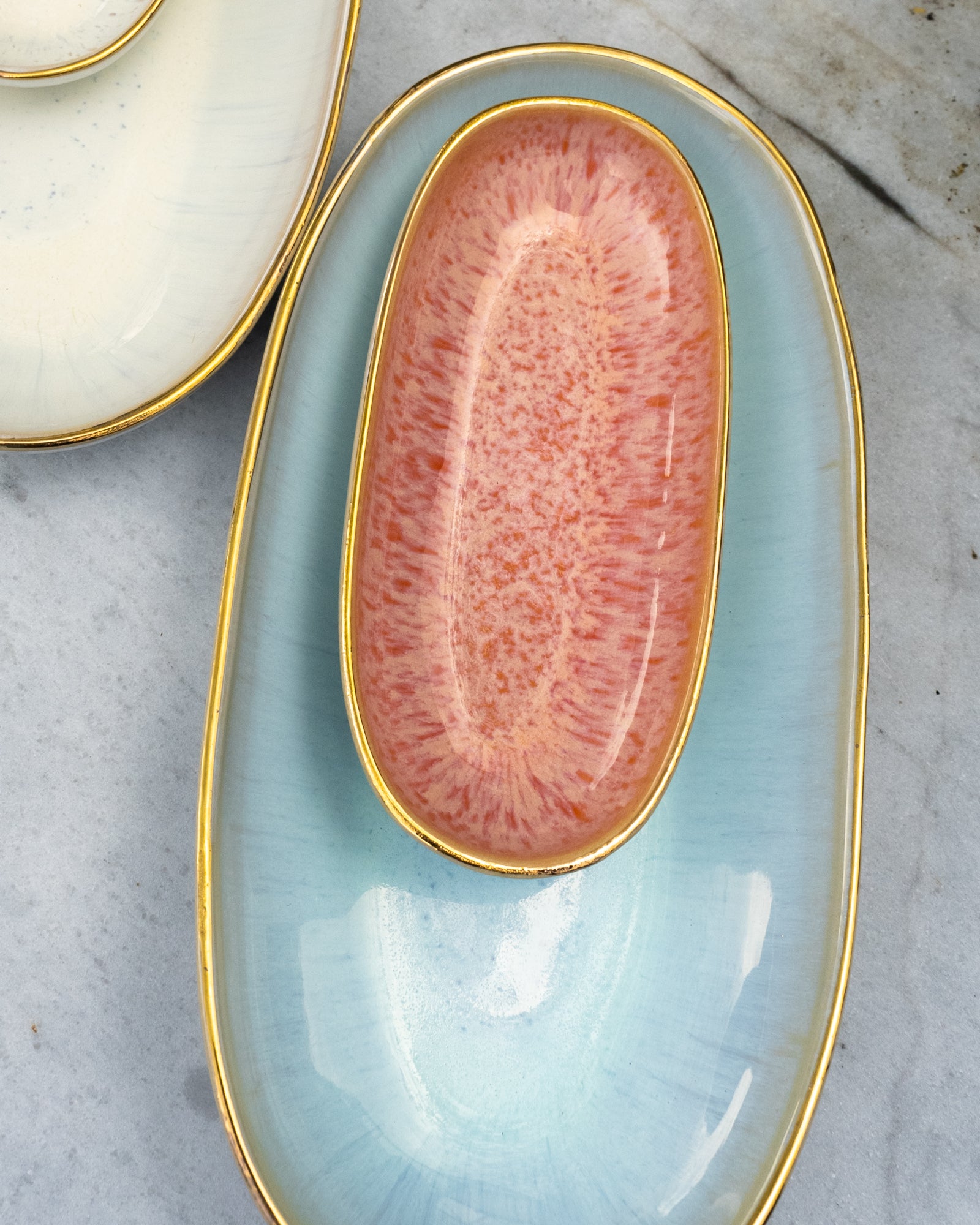 Zwei ineinander gesetzte, ovale Teller mit goldenen Rändern, der obere in Rosa, der untere in Hellblau.