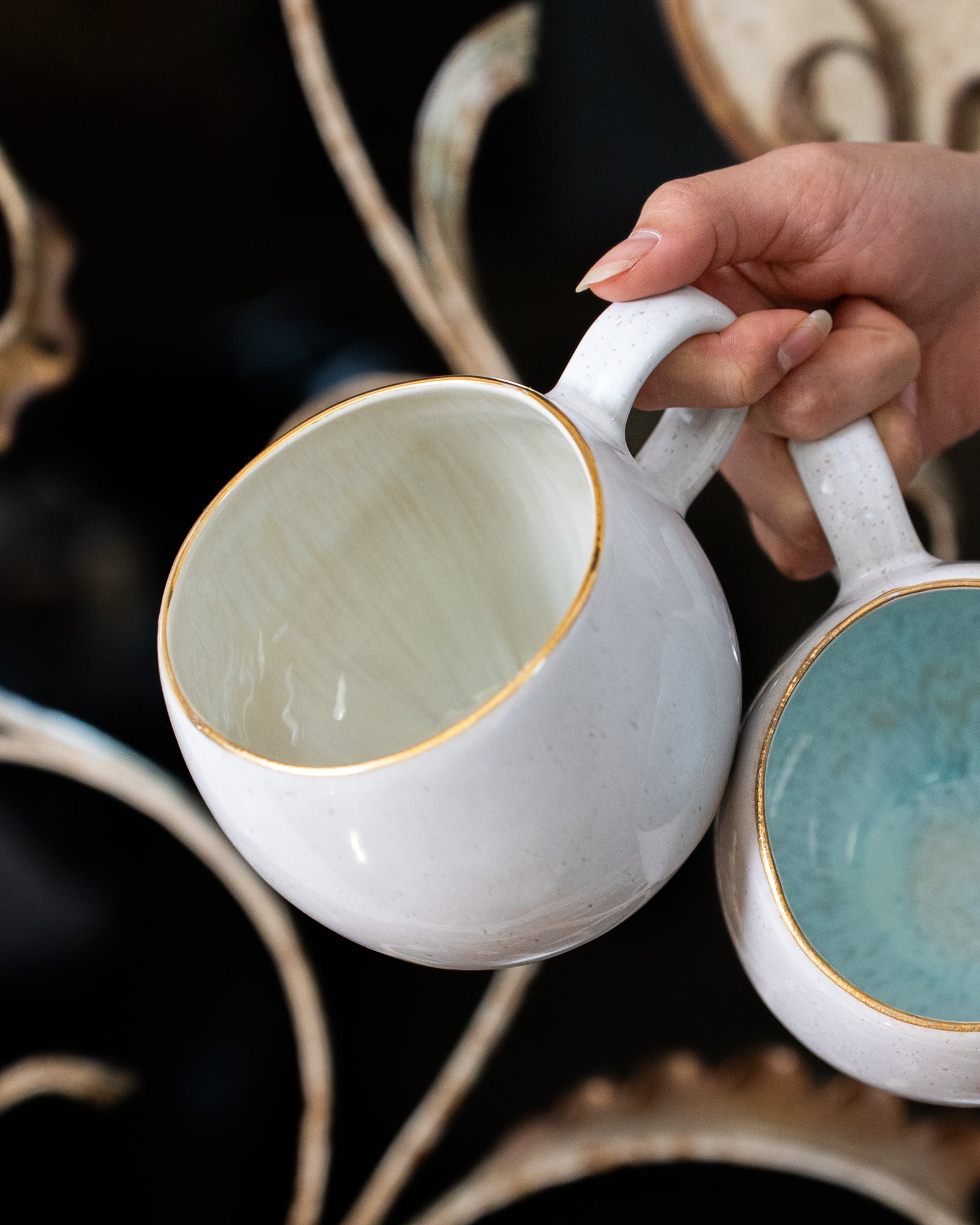 Eine Hand hält zwei weiße Keramiktassen mit goldenen Rändern, eine mit hellem und eine mit türkisfarbenem Inneren.