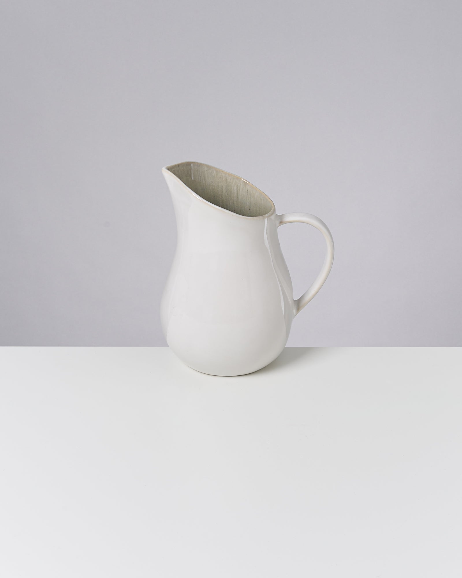 Eine weiße, keramikartige Kanne steht auf einer weißen Oberfläche vor einem grauen Hintergrund.