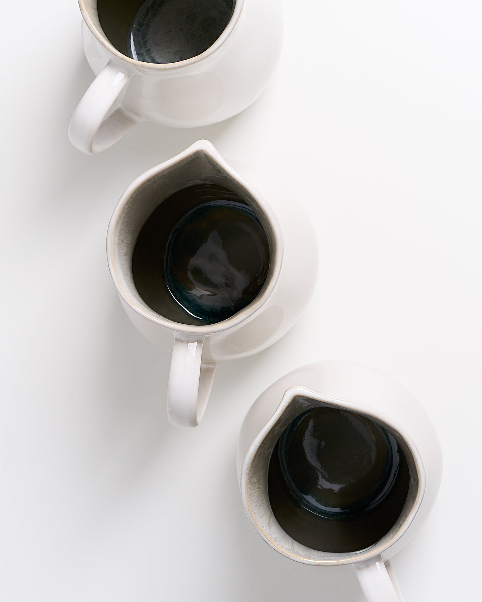 Drei weiße Keramikkrüge mit dunklem Inneren auf weißem Hintergrund von oben gesehen.