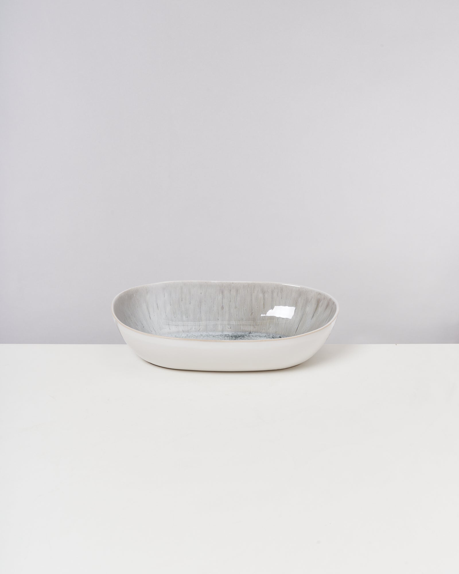 Weiße, flache, ovale Keramikschale auf einem weißen Untergrund vor einem grauen Hintergrund.